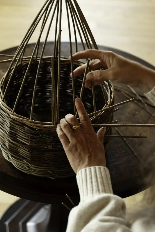 A person weaving a round wicker basket using thin wooden sticks, with their hands adjusting the structures on a dark wooden table.