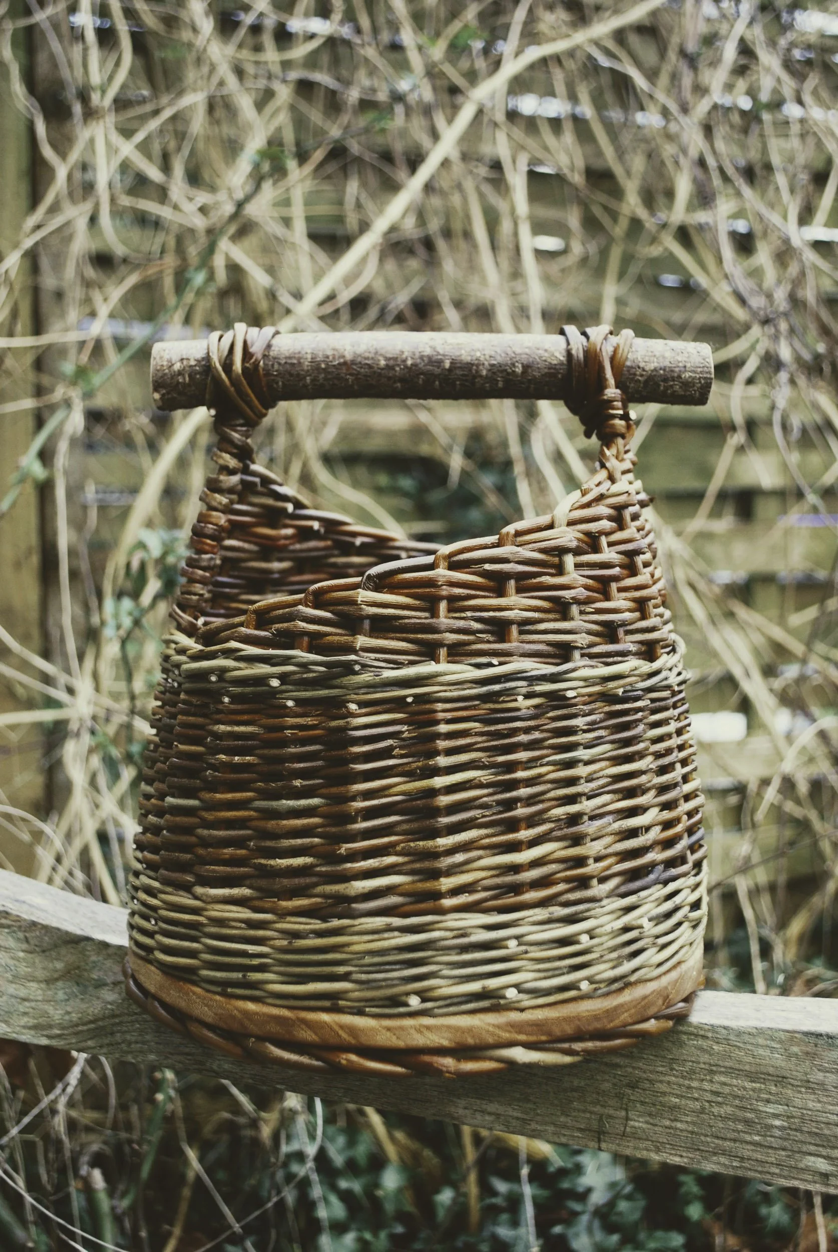 Close-up of a woven basket with a wooden handle, placed on a wooden surface, with a background of a lattice fence and dried vines.