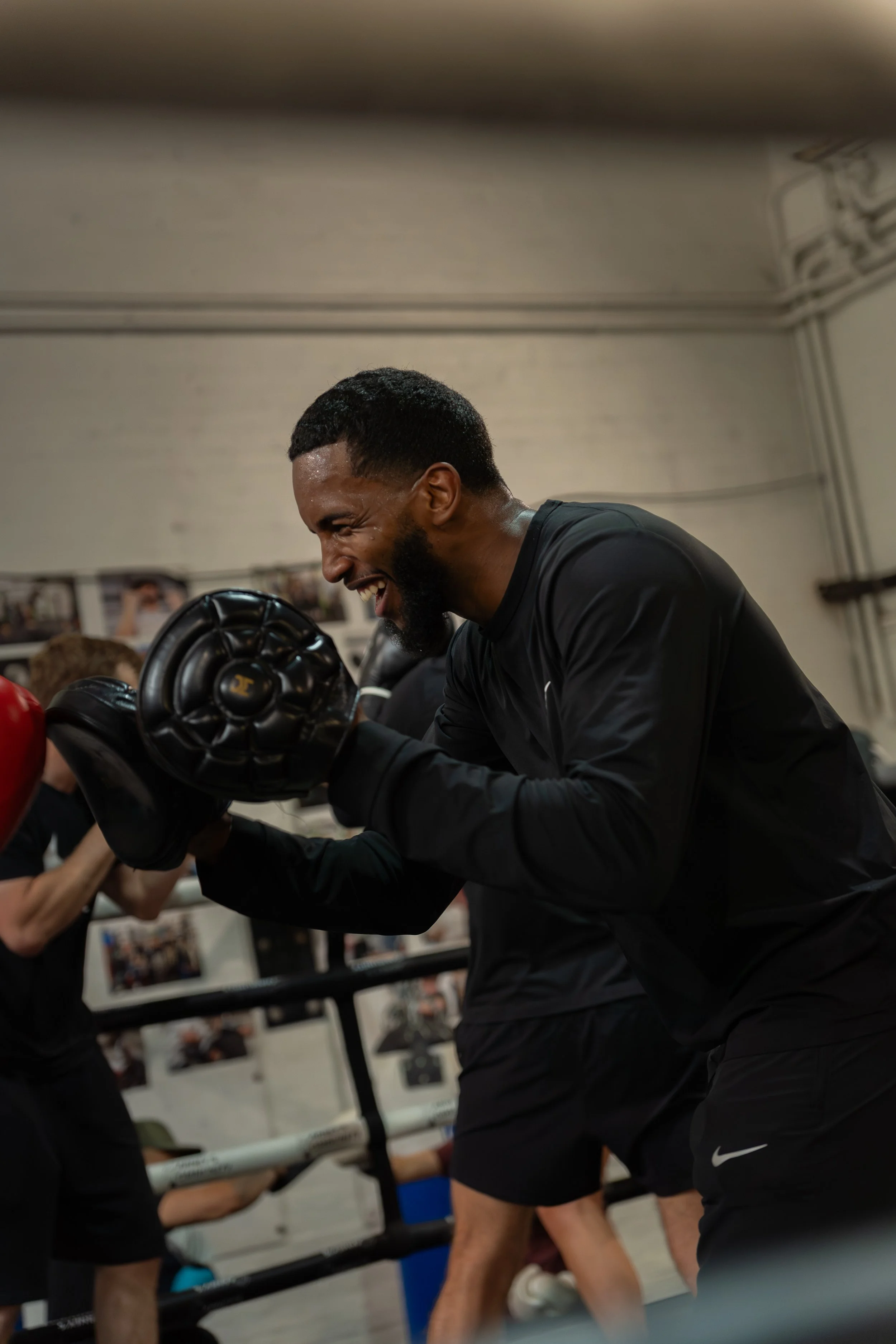 A man smiling and wearing boxing gloves during a training session or sparring with another boxer in a gym.