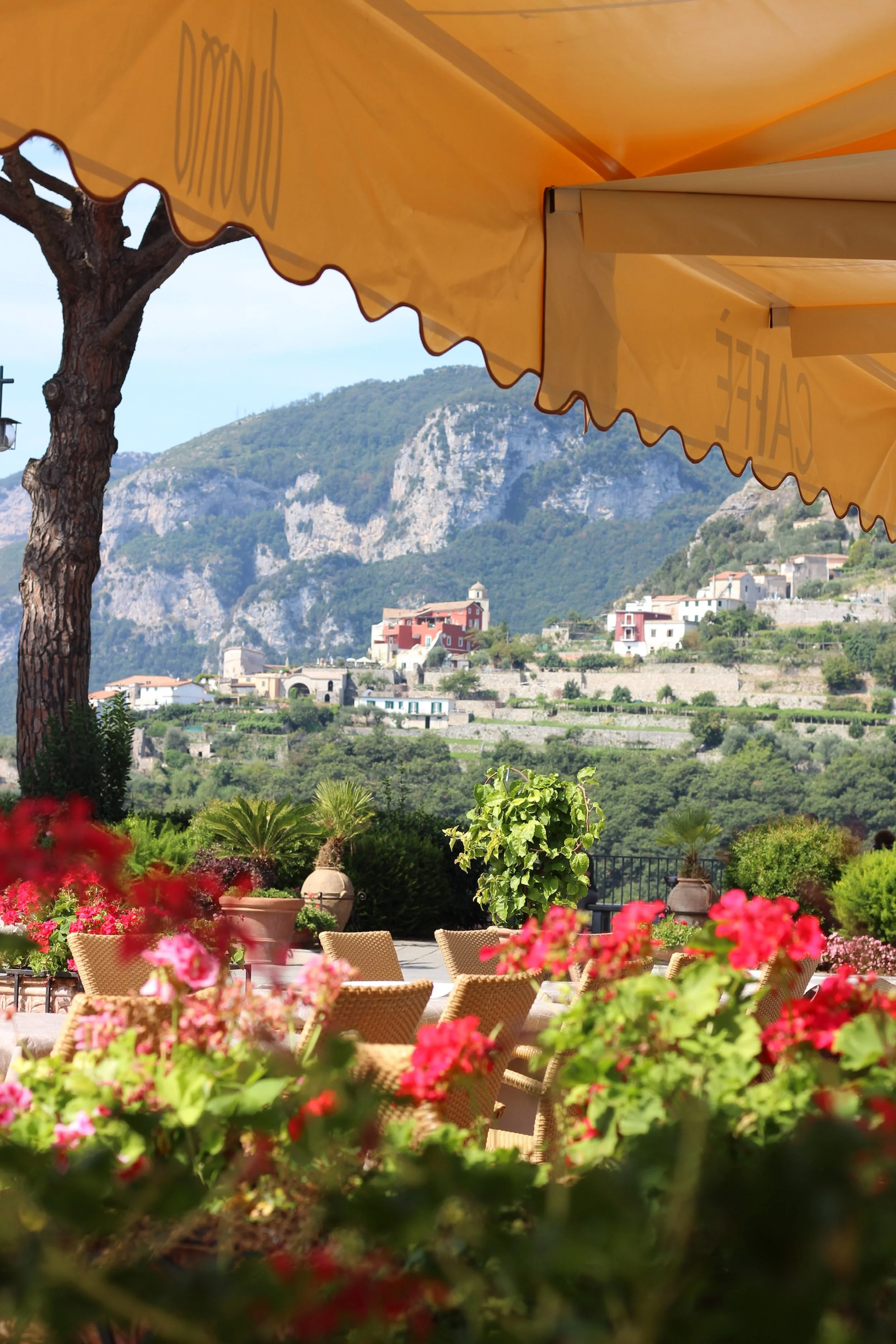 Outdoor cafe with wicker chairs and vibrant pink flowers, overlooking a hillside town with houses and lush green trees under a yellow awning in Italy.