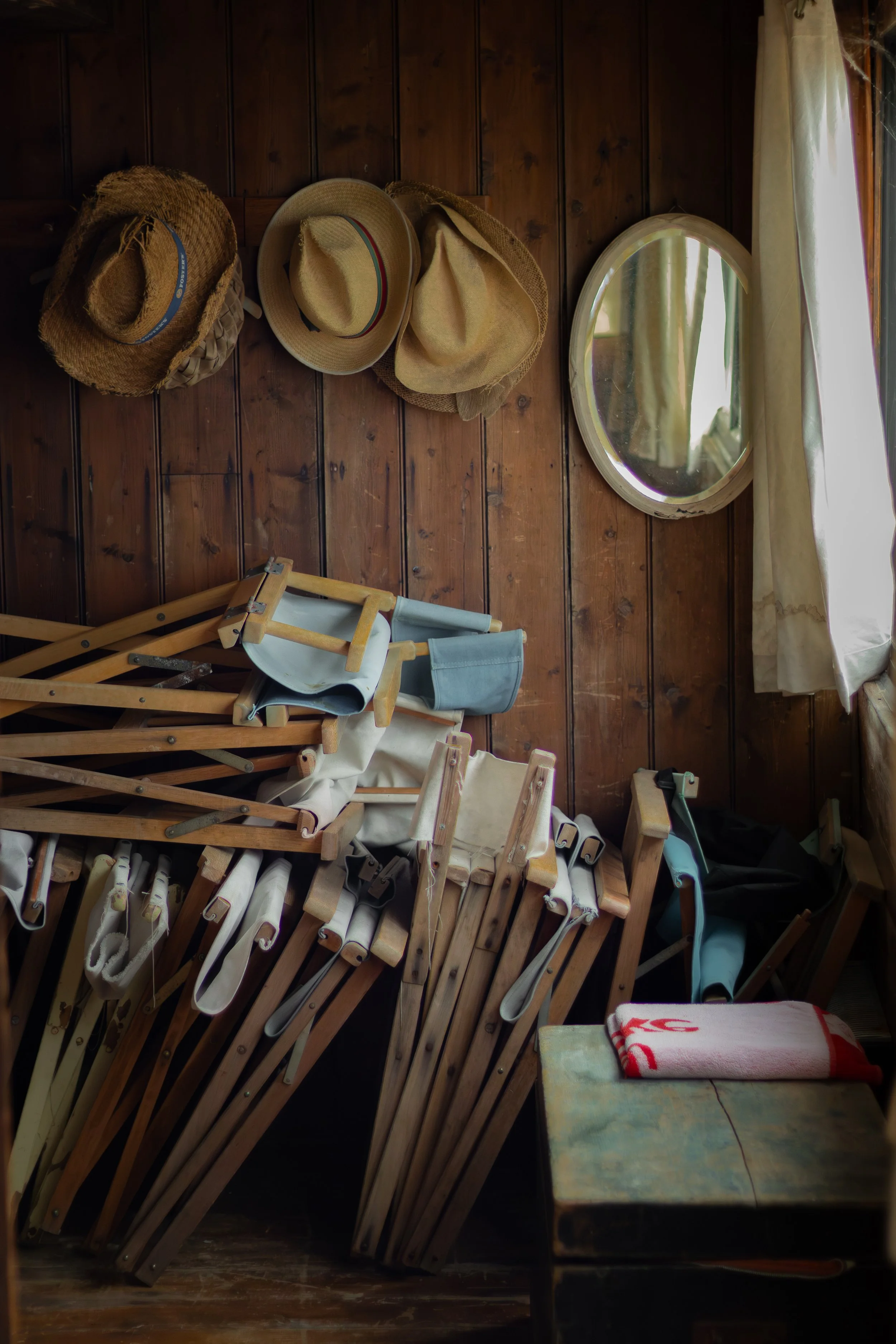 A collection of straw hats hanging on a wooden wall, with a mirror and a window with curtains in the background, and a table with a towel and various objects in the foreground.