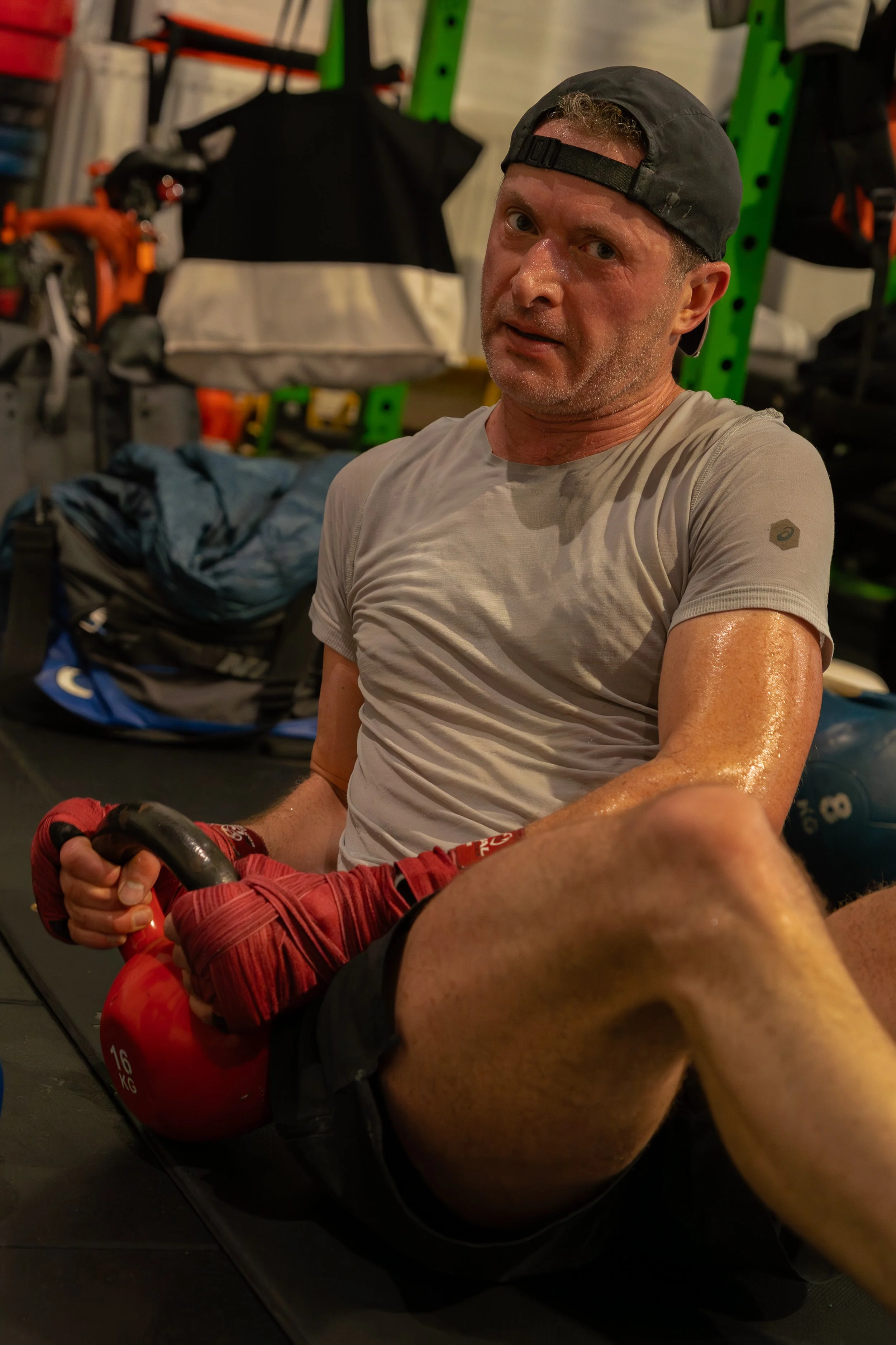 Man sweating and sitting on the floor in a gym, holding a red kettlebell. The gym has green weight racks and bags in the background.