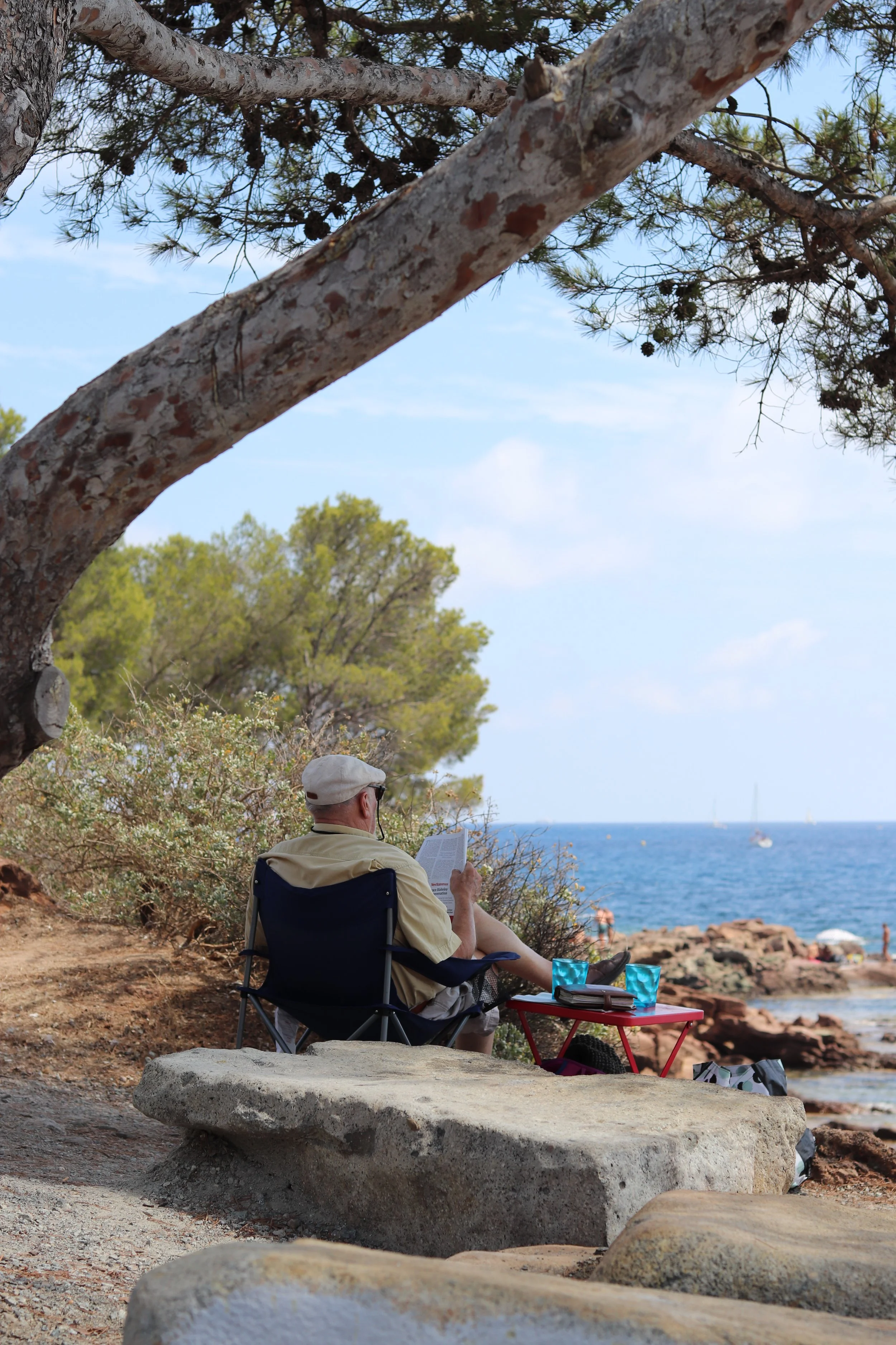 An elderly man sitting in a camping chair under a tree near the coast in France, reading a book with two glasses and a small red table nearby, overlooking the ocean with rocks and sailboats in the distance.