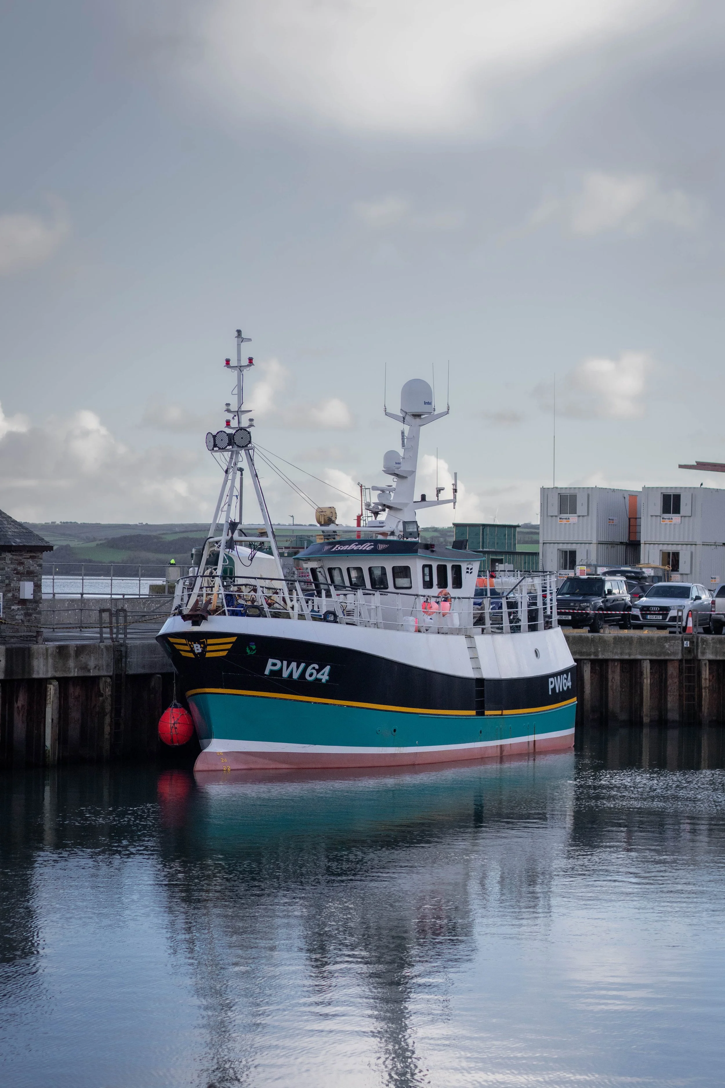 A fishing boat docked at a harbor on calm water, with a cloudy sky overhead in Padstow, Cornwall, United Kingdom.