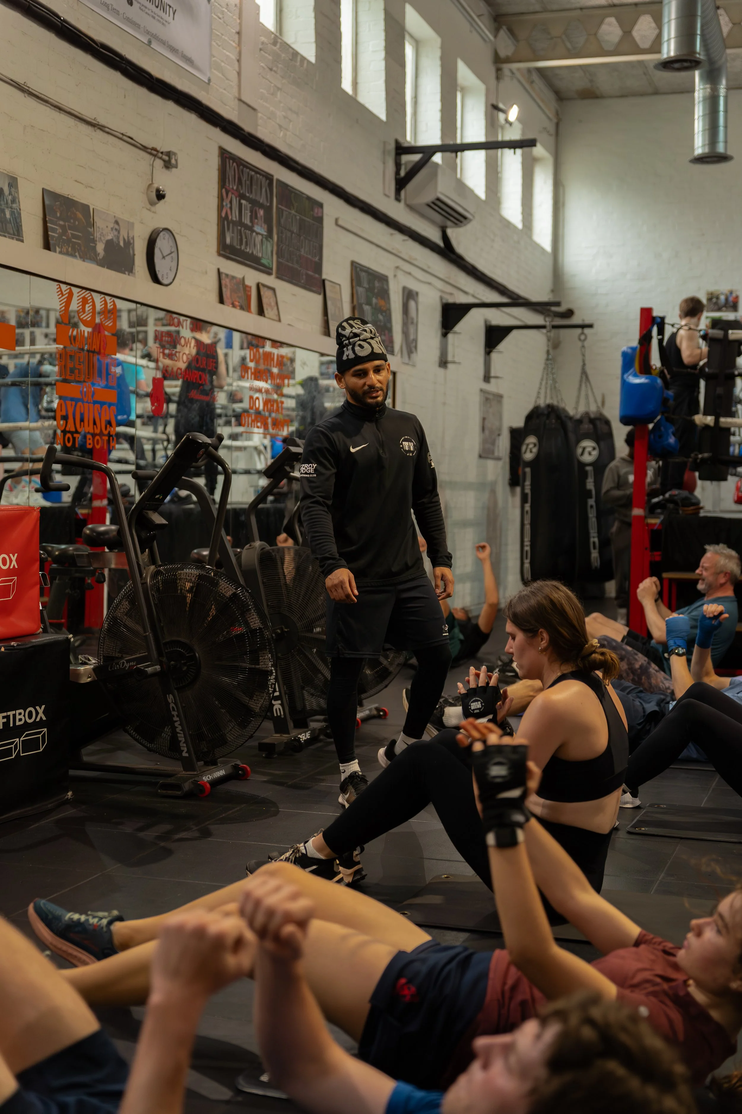 Group fitness class in a gym with people stretching and exercising, some seated and some lying on the floor, with fitness equipment and motivational signs on the walls.