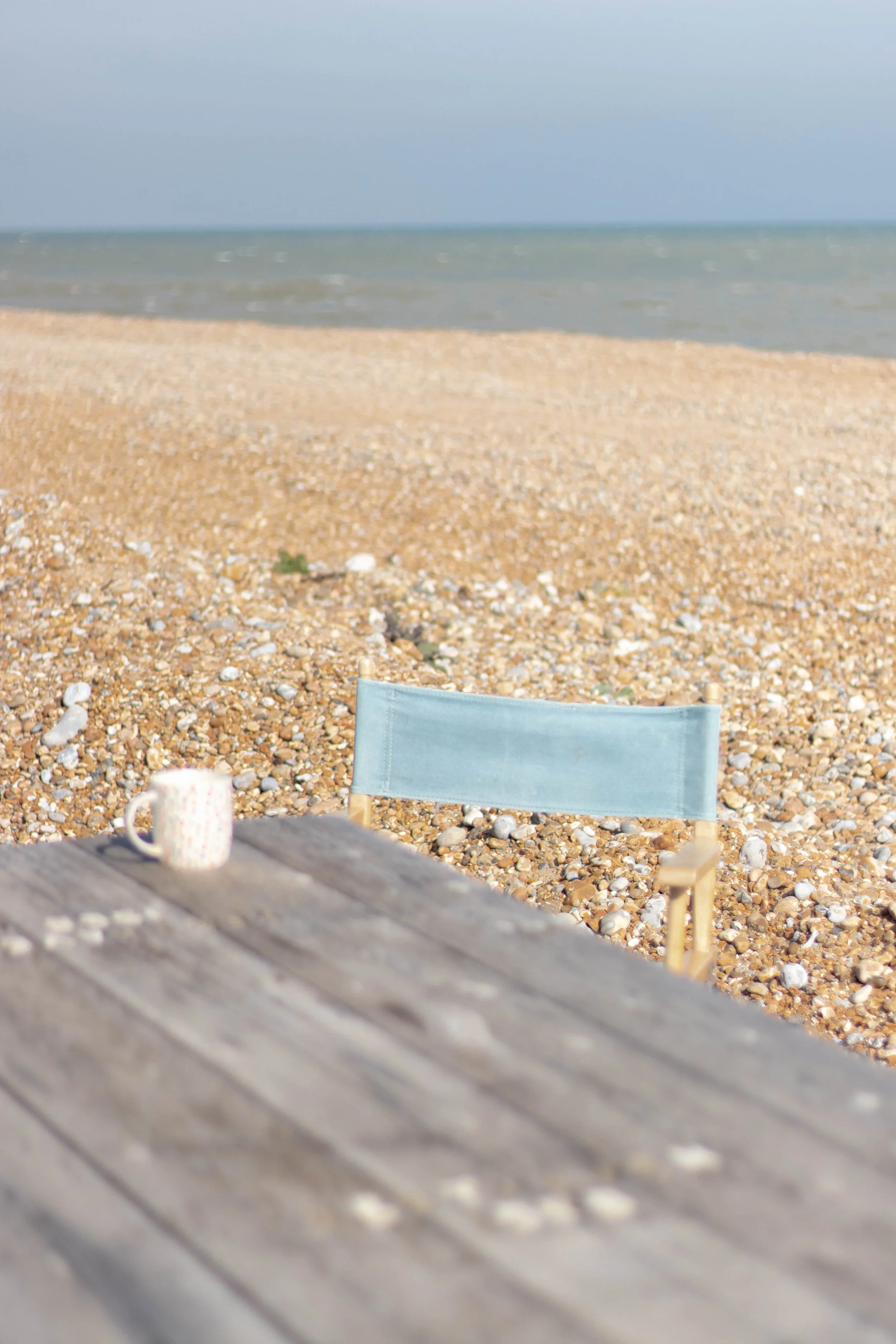 A beach scene with a weathered wooden table, a ceramic mug, and a folding chair on a pebbled shore overlooking the ocean under a cloudy sky in Norman's Bay, United Kingdom.