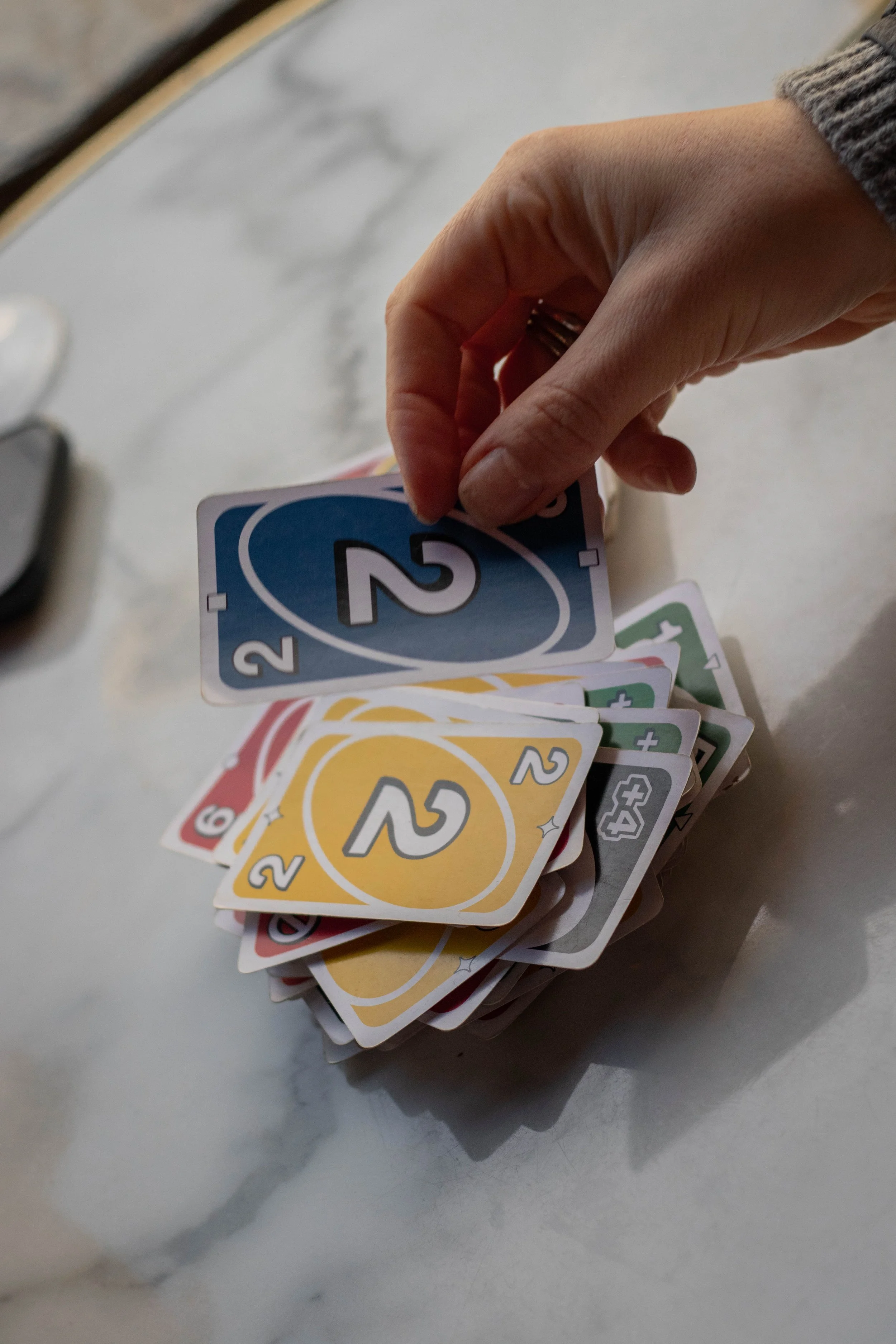 A hand holding a blue '2' card above a stack of various coloured UNO cards on a marble surface.