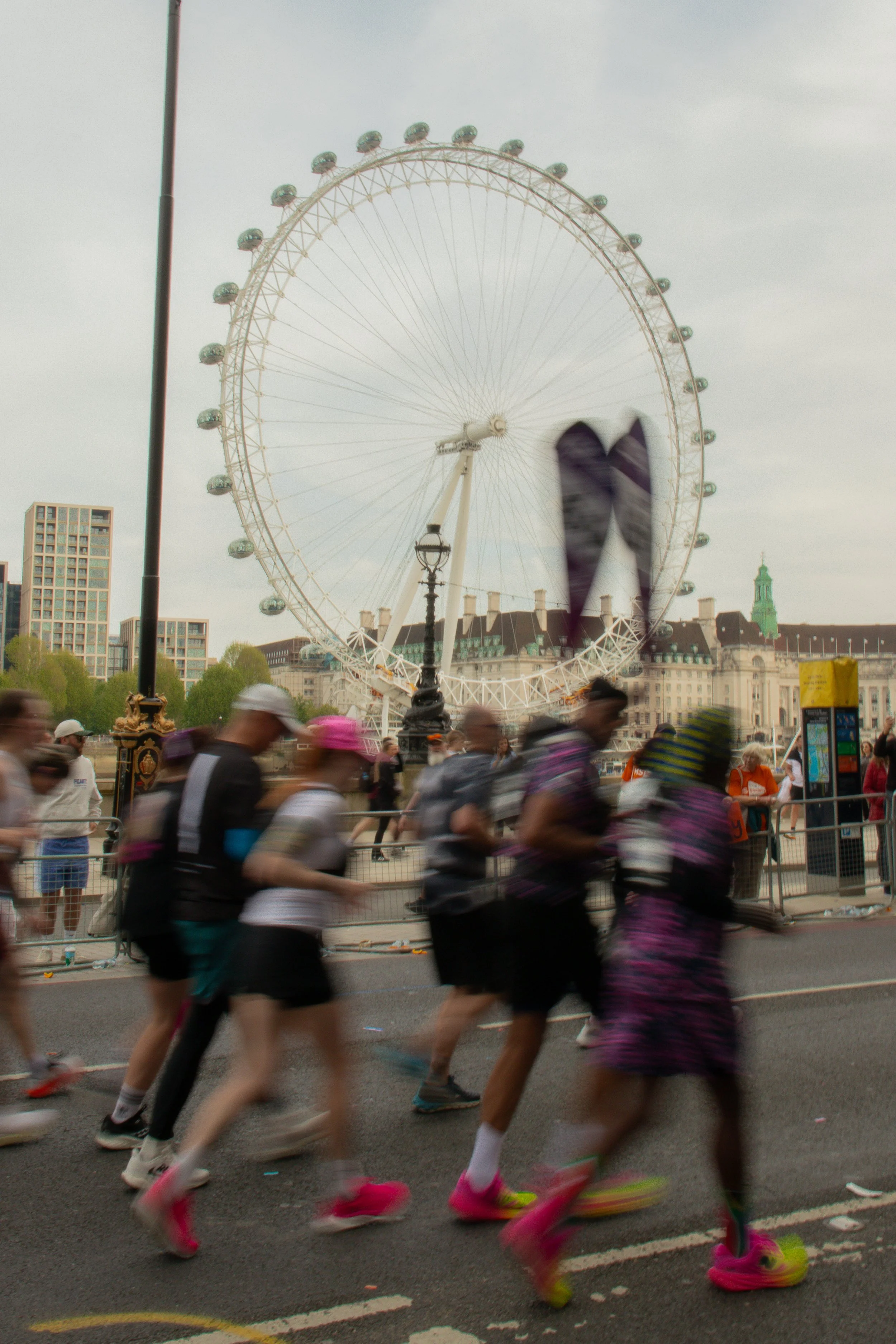 A group of runners participating in the London Marathon, with the London Eye, buildings, and a cloudy sky in the background.
