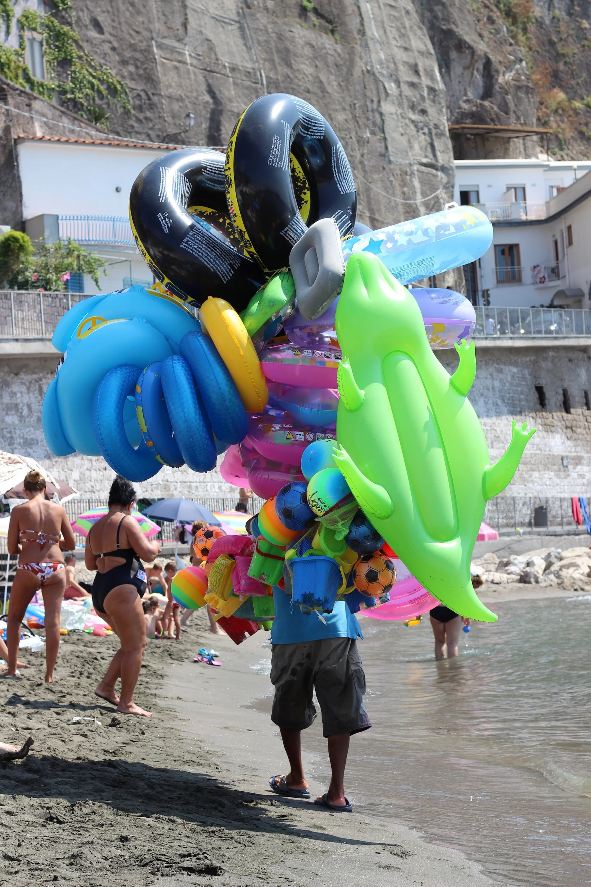 Beach scene in Italy with people, colourful inflatable pool toys, and a person carrying a large bundle of inflatables near the shoreline.