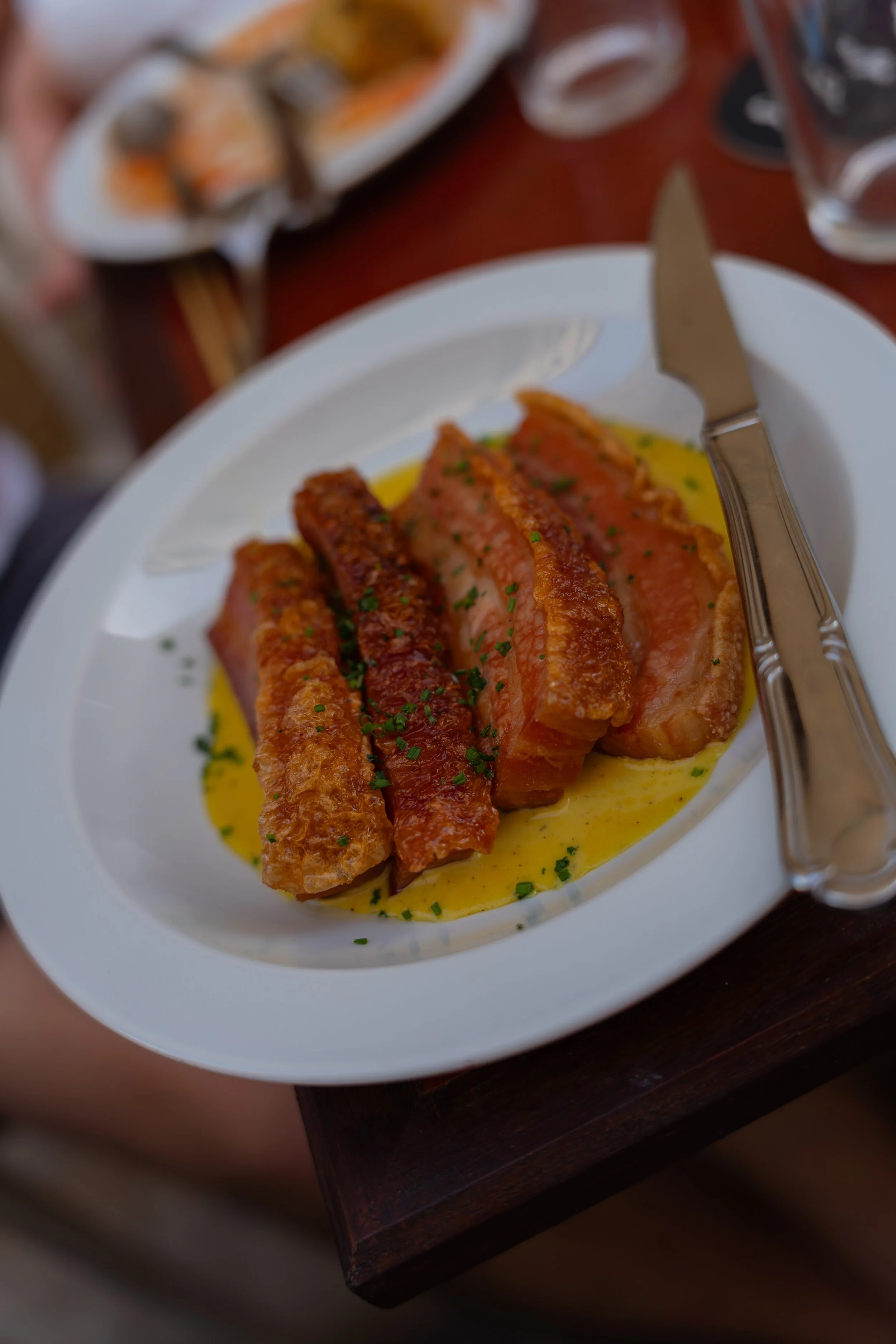 Close-up of a white plate with slices of cooked pork covered in herbs, served with yellow sauce at the La Nouvelle Famille restaurant in Majorca, Spain. In the background, part of another dish and utensils are visible on a wooden table.