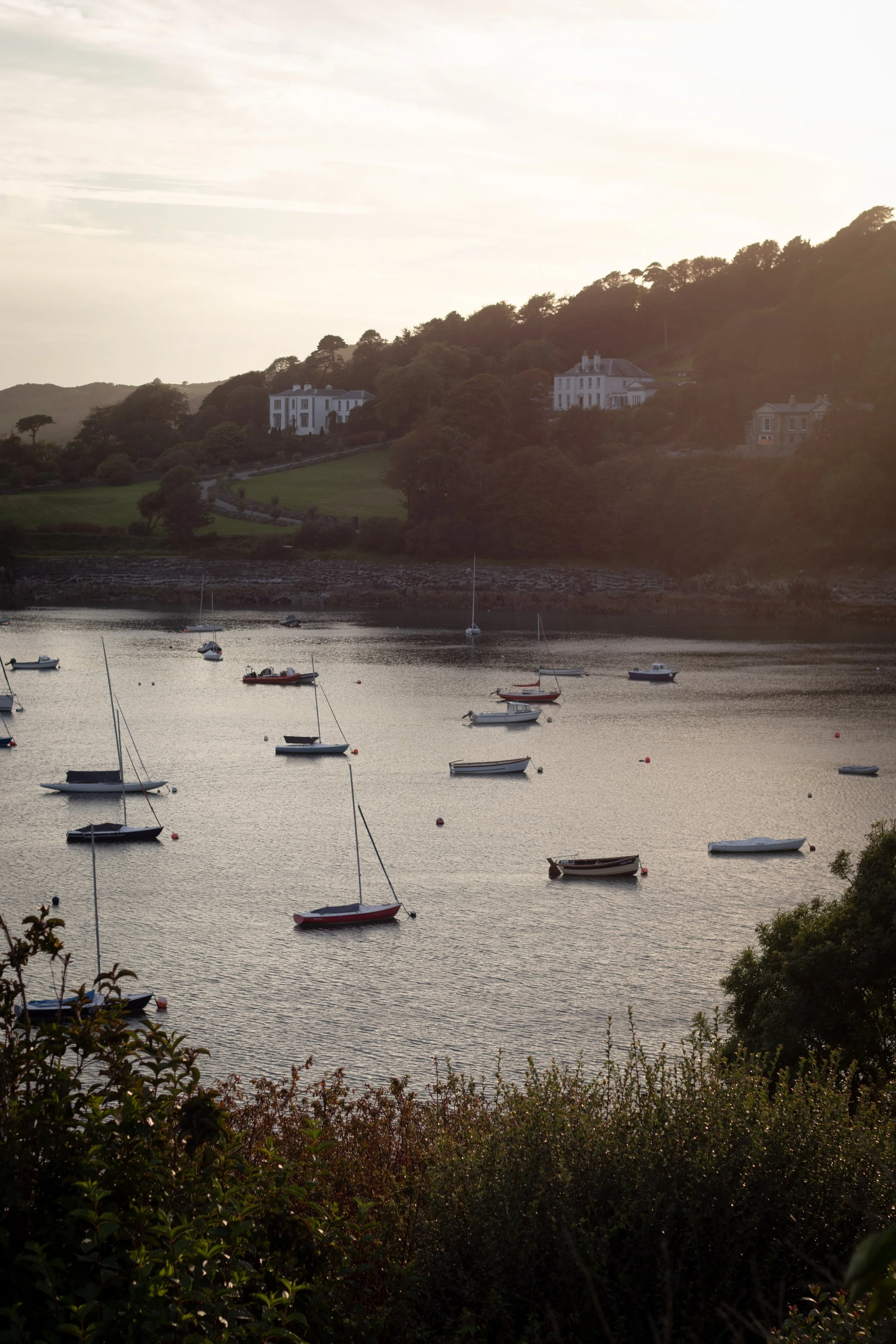 Scenic view of a harbor with sailboats floating on calm water, overlooking green hills with large white houses at sunset in Ireland.