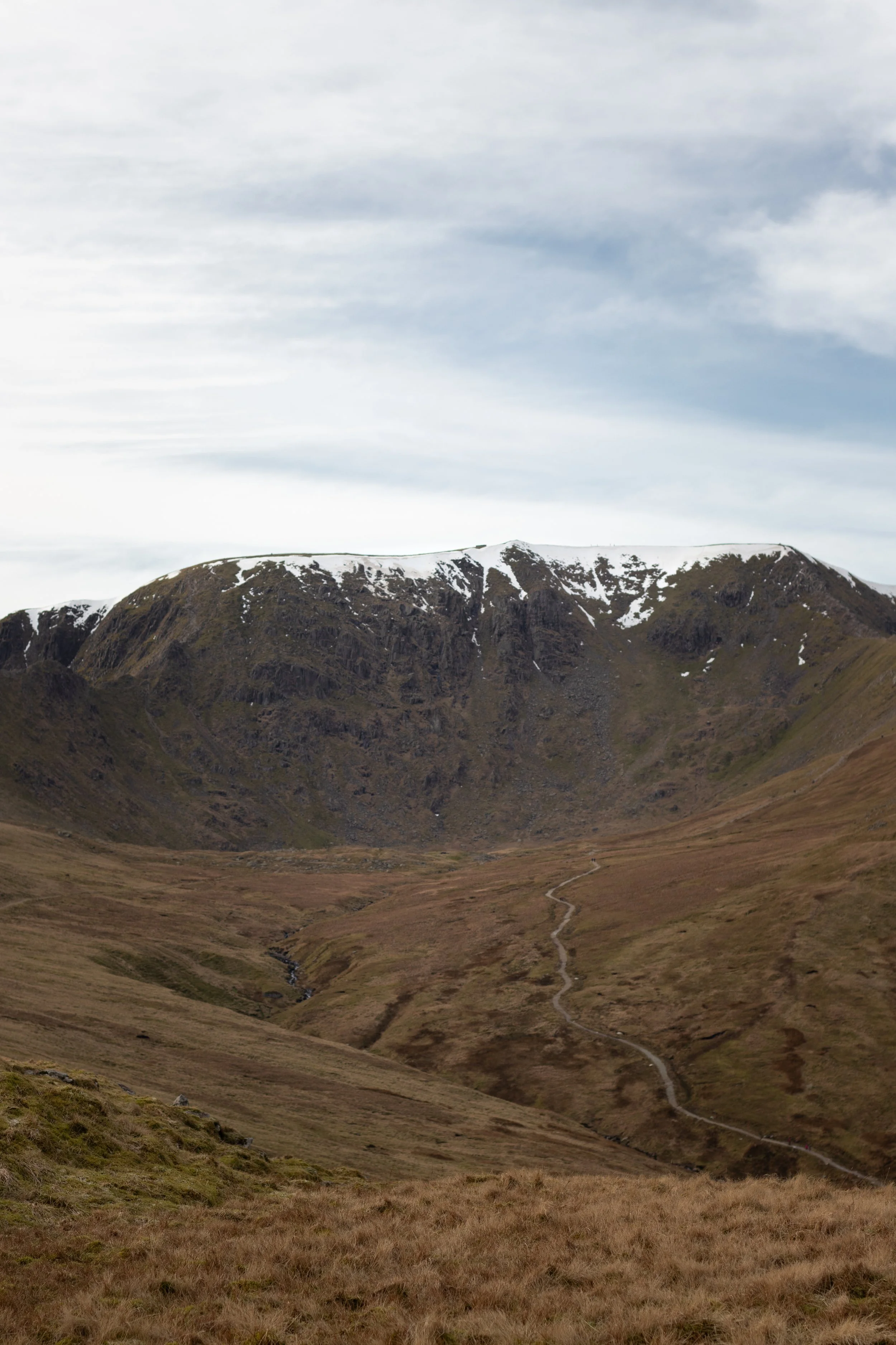 A mountainous landscape with snow patches on the peak and a winding trail through grassy slopes under a cloudy sky in The Lake District, United Kingdom.