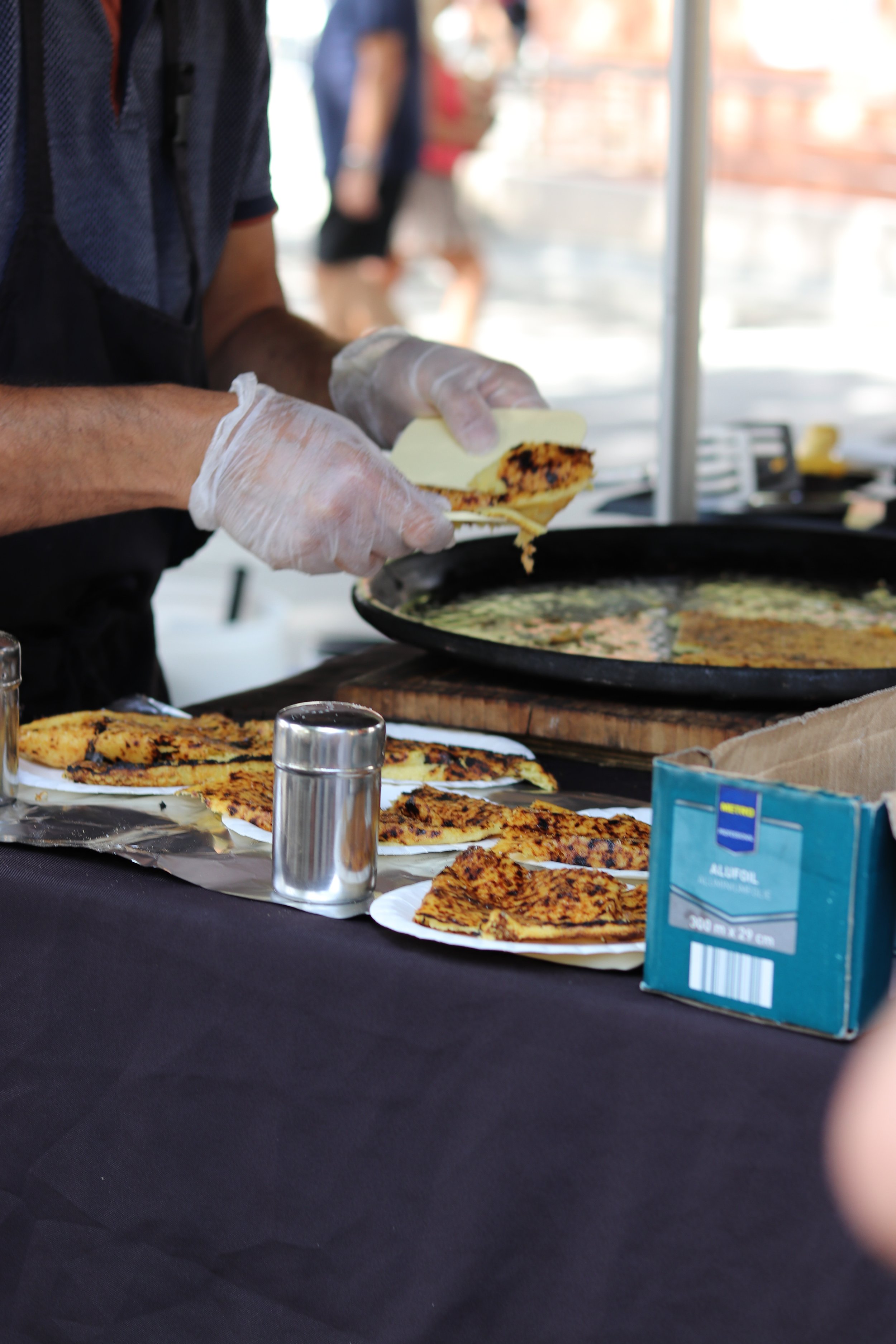 Person preparing grilled quesadillas at a food stall in France, with several cooked quesadilla slices on the table, a metal salt shaker, and a box of aluminium foil.