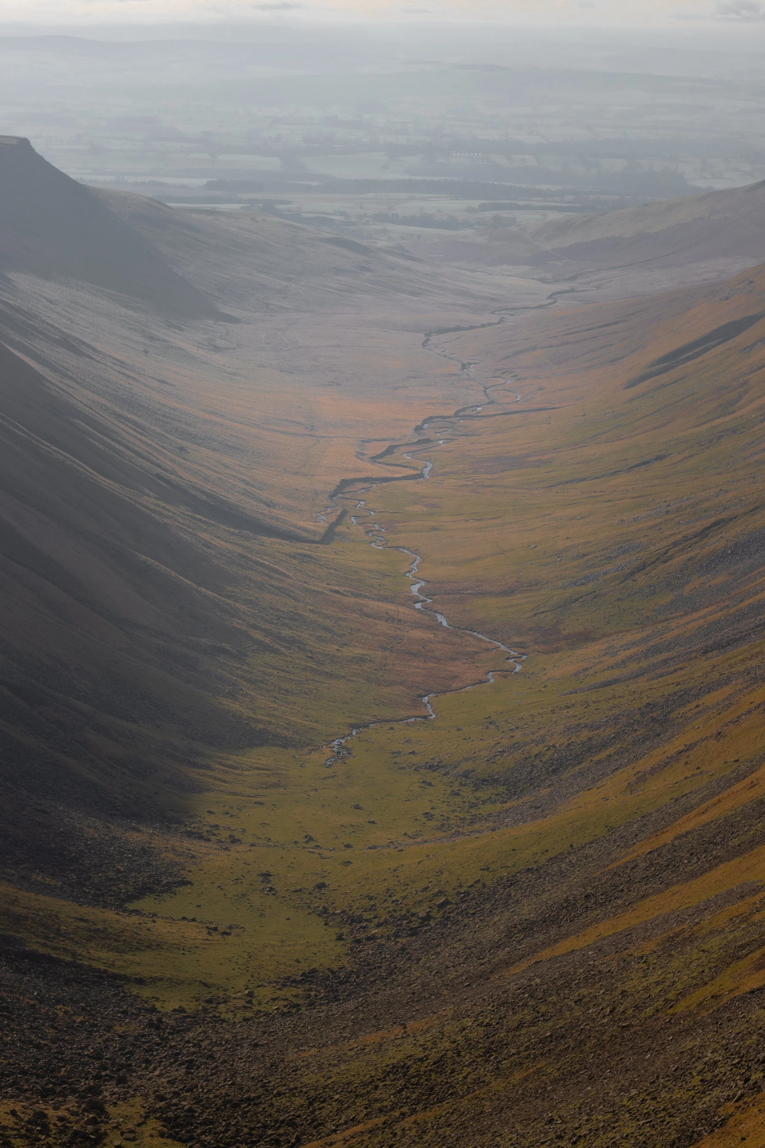 A bird's eye view of a green and brown valley with a small meandering stream running through it, surrounded by steep mountains in Cumbria, United Kingdom.