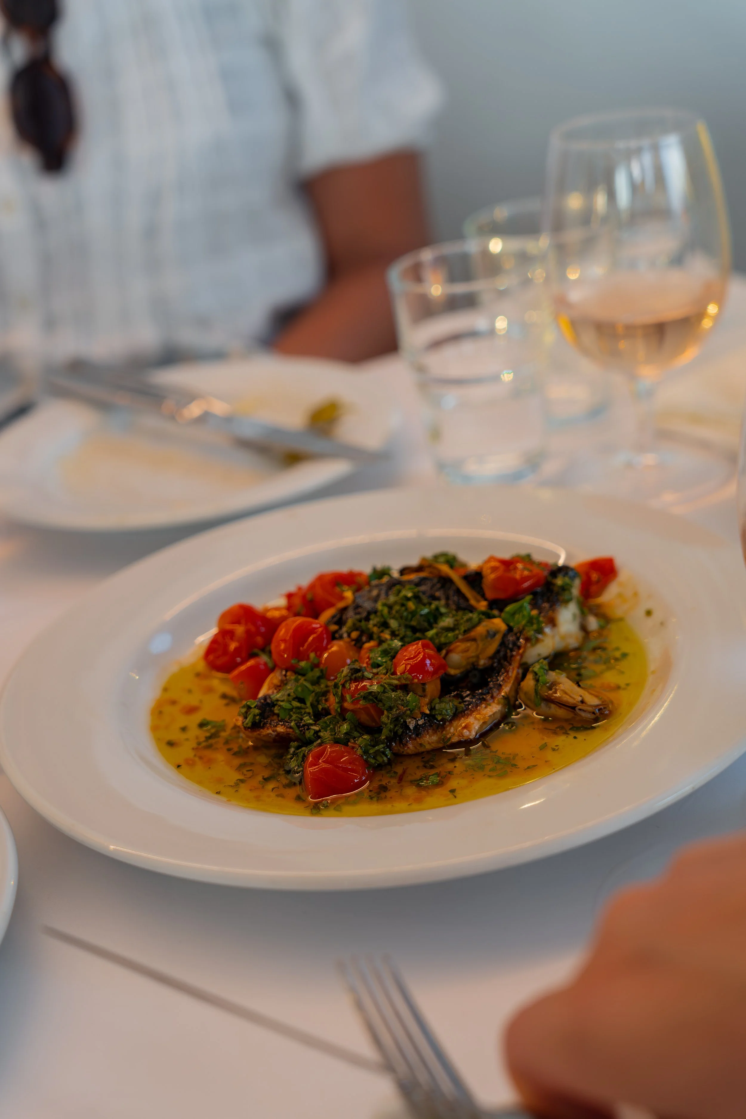 A plate of cooked fish topped with cherry tomatoes and herbs, with a pool of sauce underneath, on a white tabl. In the background, there are three glasses of rosé wine, a person's torso, now blurred, and a table setting with cutlery and a folded napk