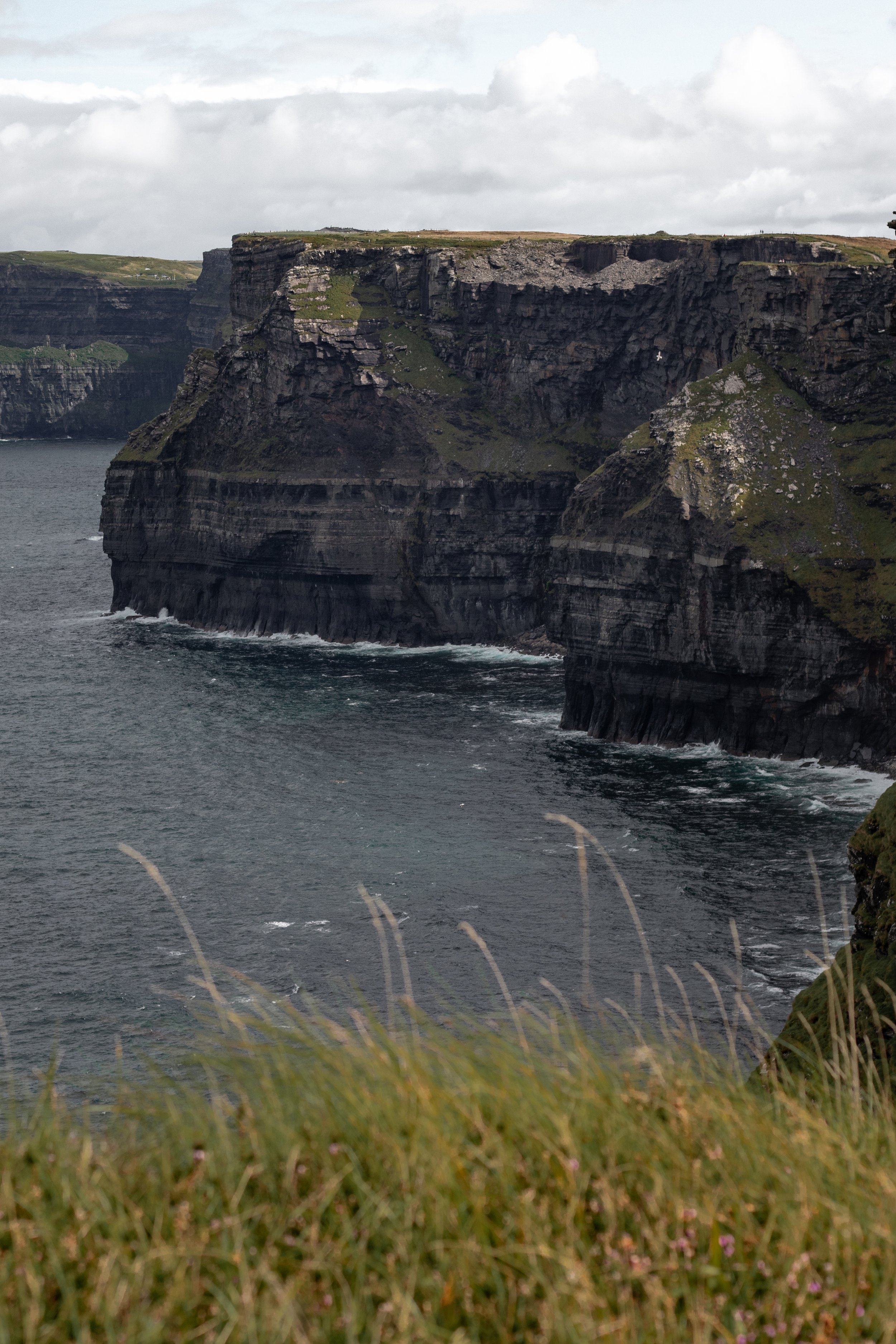 Cliffs along a rocky coastline with grassy foreground and cloudy sky in Ireland.