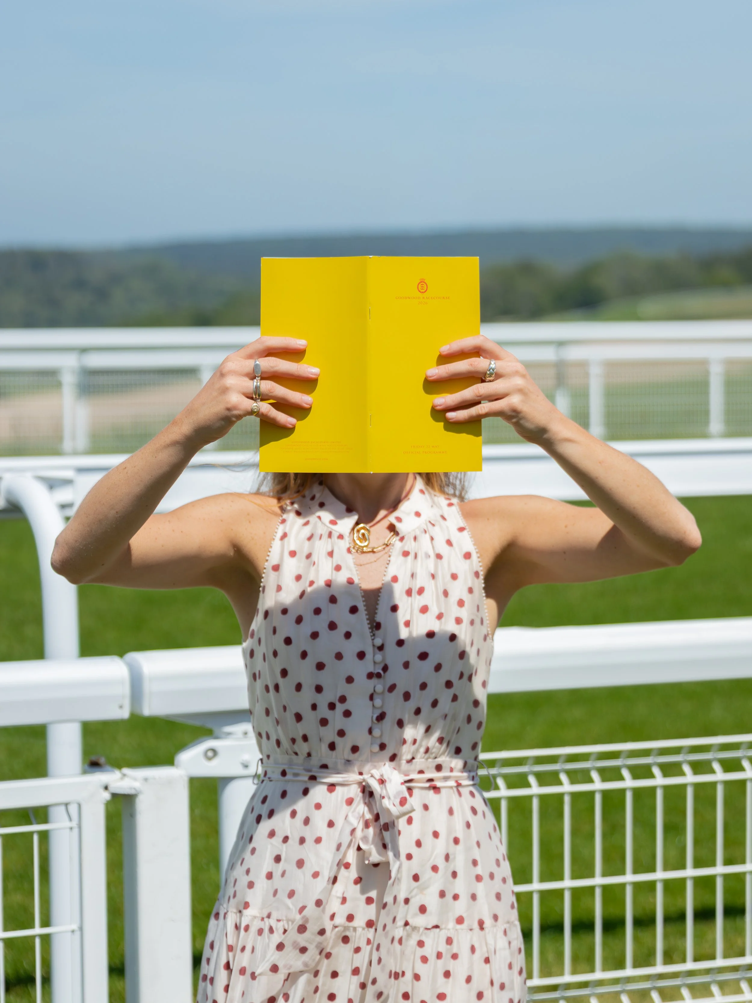 A person wearing a polka dot Phase Eight dress is holding a yellow book in front of their face, standing trackside at Goodwood Racecourse with the white fence track fence and green landscape in the background.