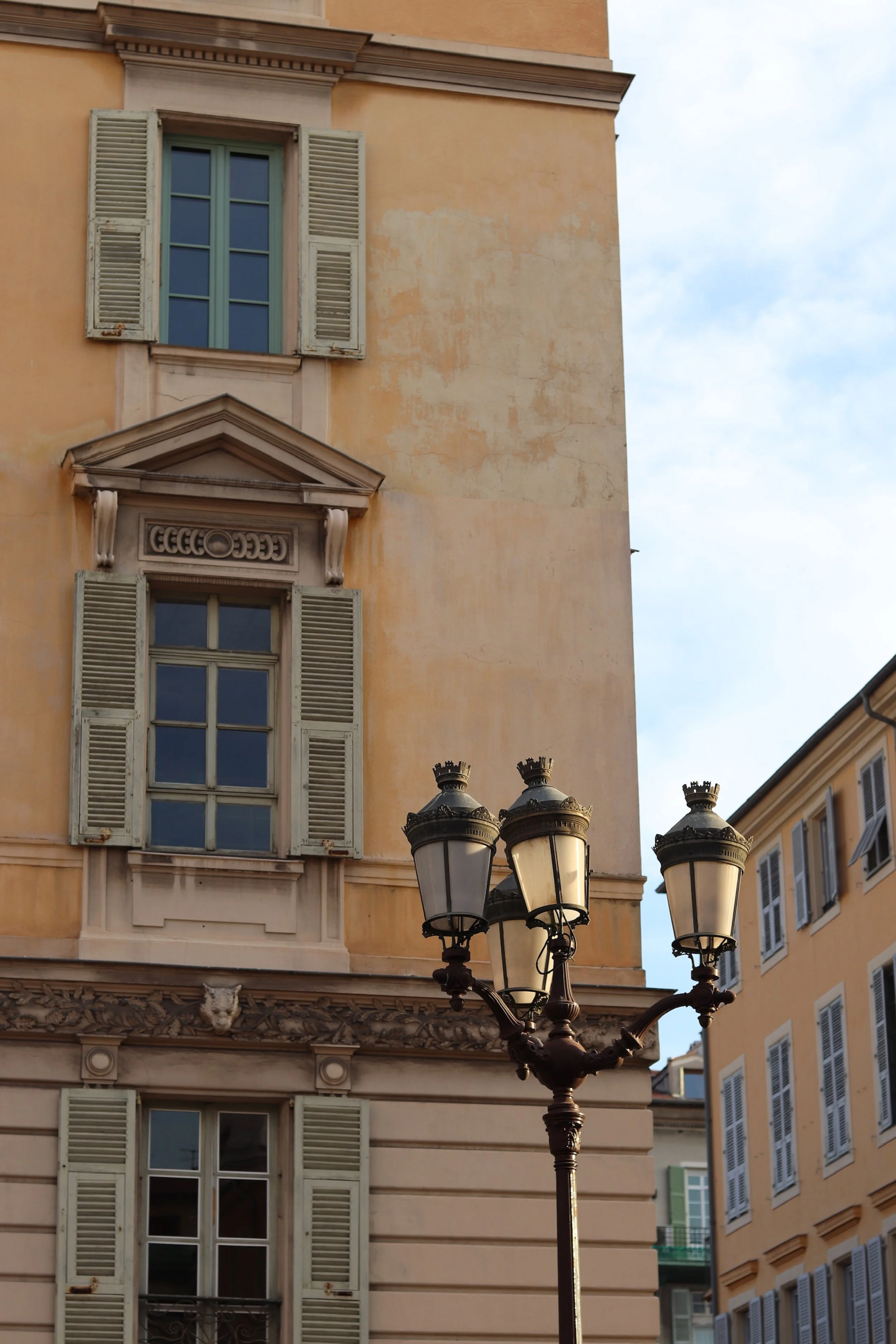 Historic building in France with yellow walls, ornate architectural details above windows, open shutters, and a vintage street lamp in the foreground.