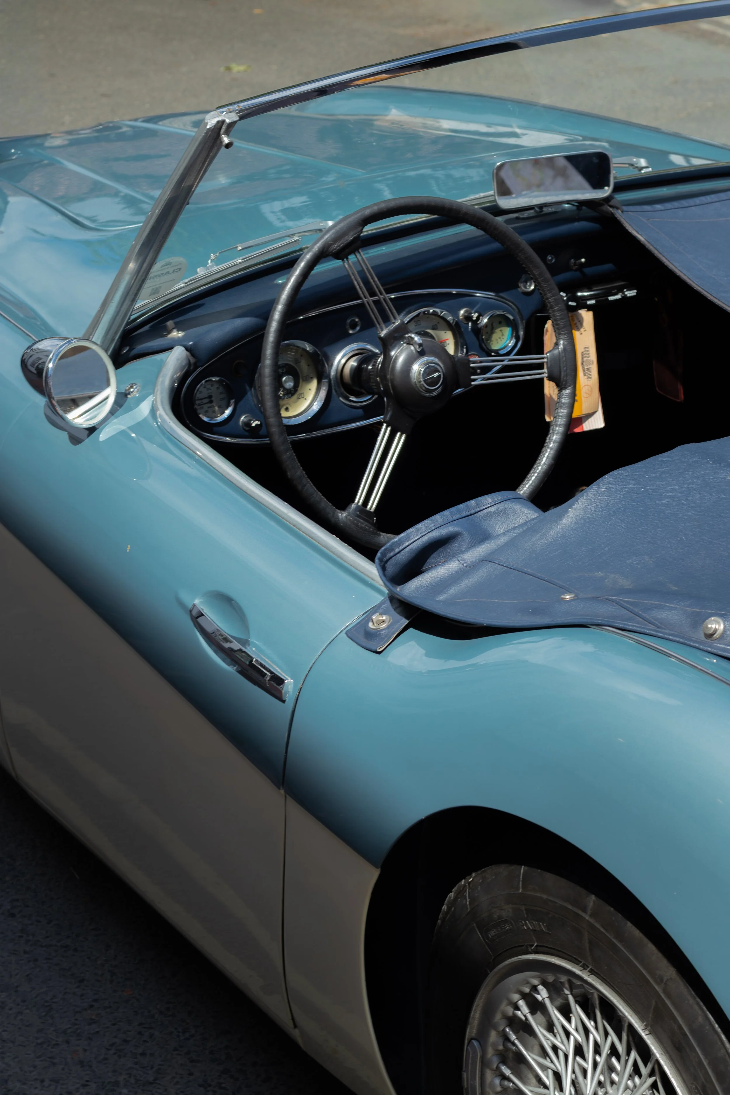 Close-up of a vintage convertible car's dashboard and front wheel, featuring classic gauges, a steering wheel, and a mirror.