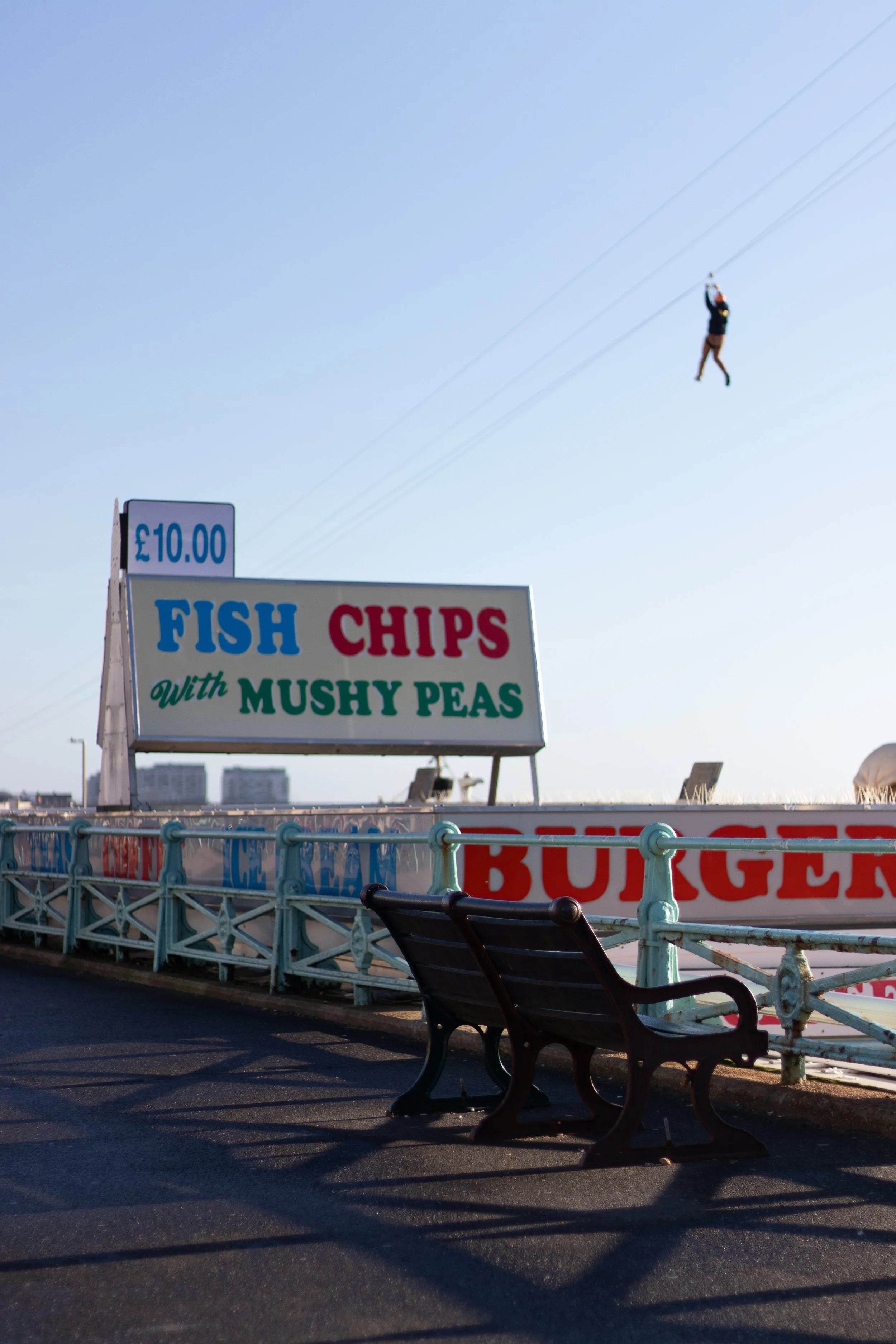 Seafront promenade in Brighton with a sign for fish and chips with mushy peas, a bench, and a person zip-lining in the sky.