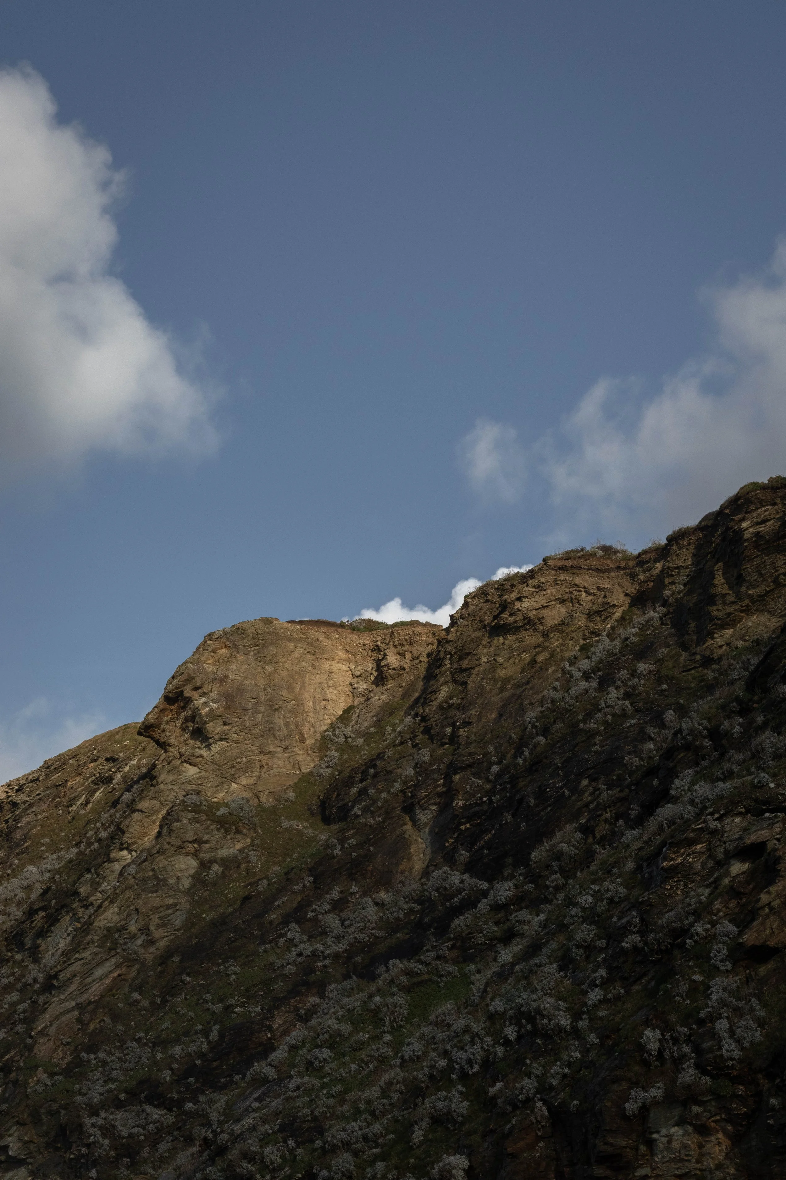 A mountain with rugged rocks and patches of vegetation against a sky with a few clouds in Cornwall, United Kingdom.