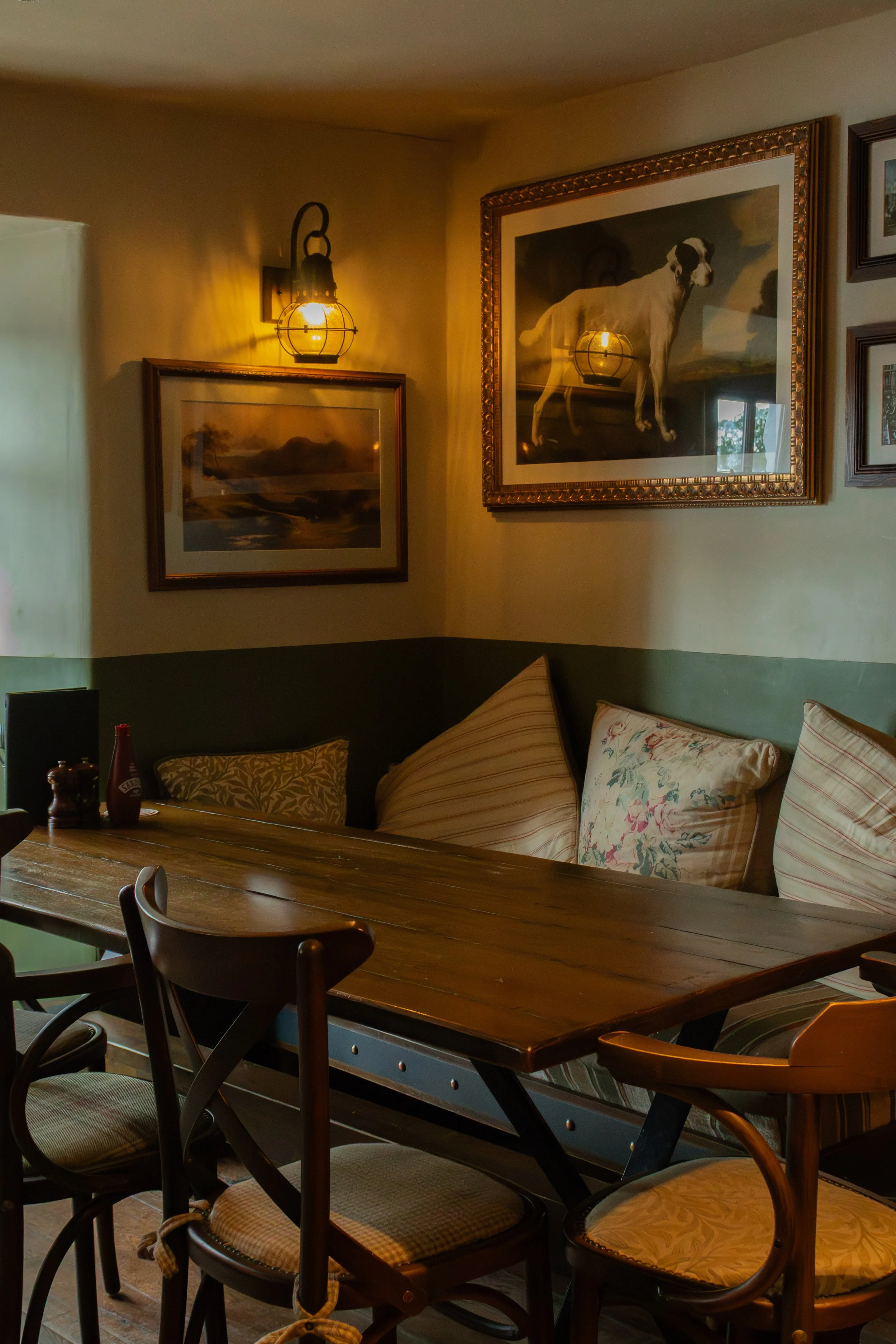 Interior of a cozy dining area with a wooden table and chairs, cushions on a bench, framed artwork on the wall, and warm lighting from a decorative wall sconce.