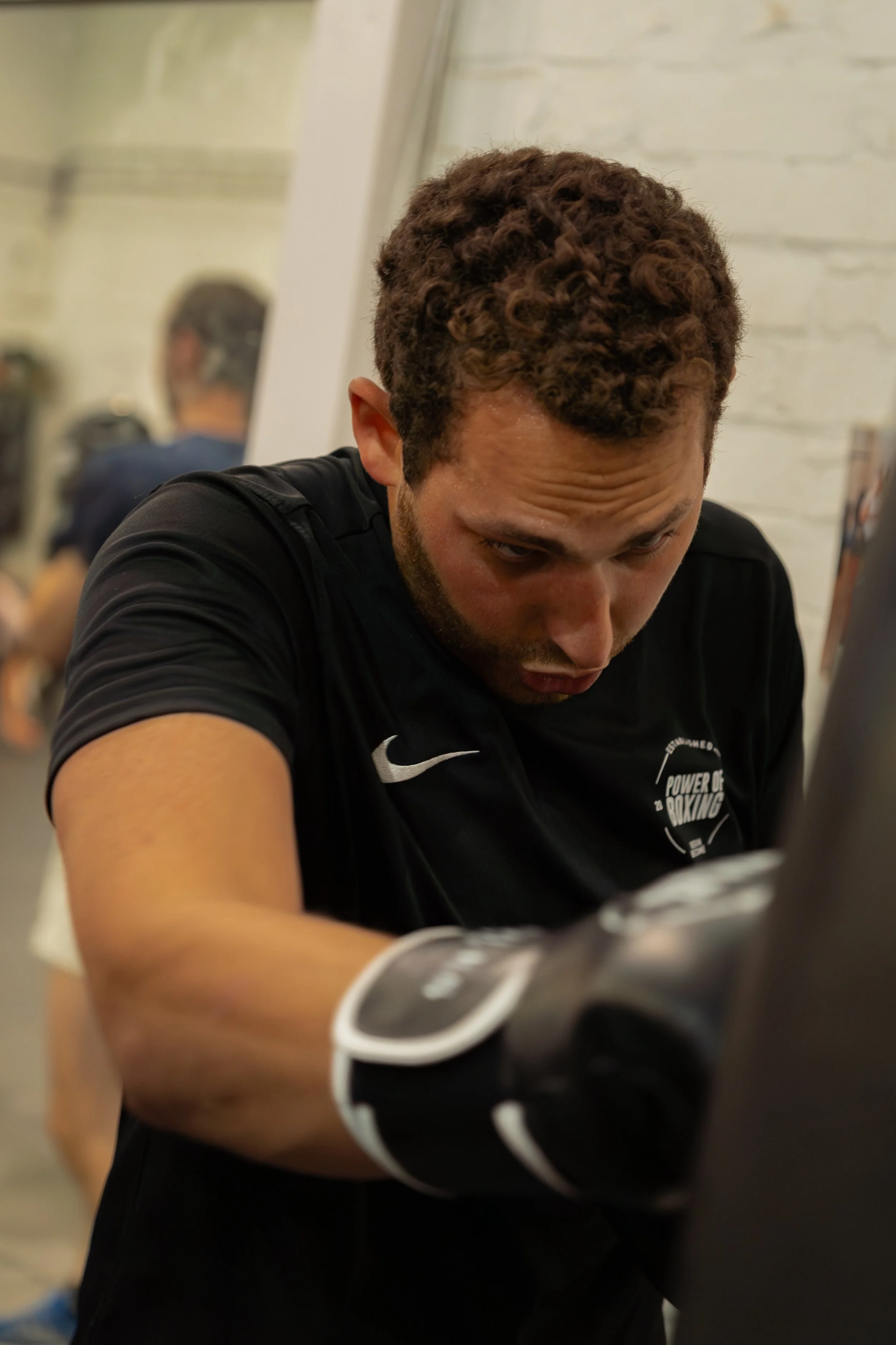 A man with curly brown hair concentrating while wearing a black athletic shirt with a white Nike logo and black gloves, in an indoor setting with blurred background.