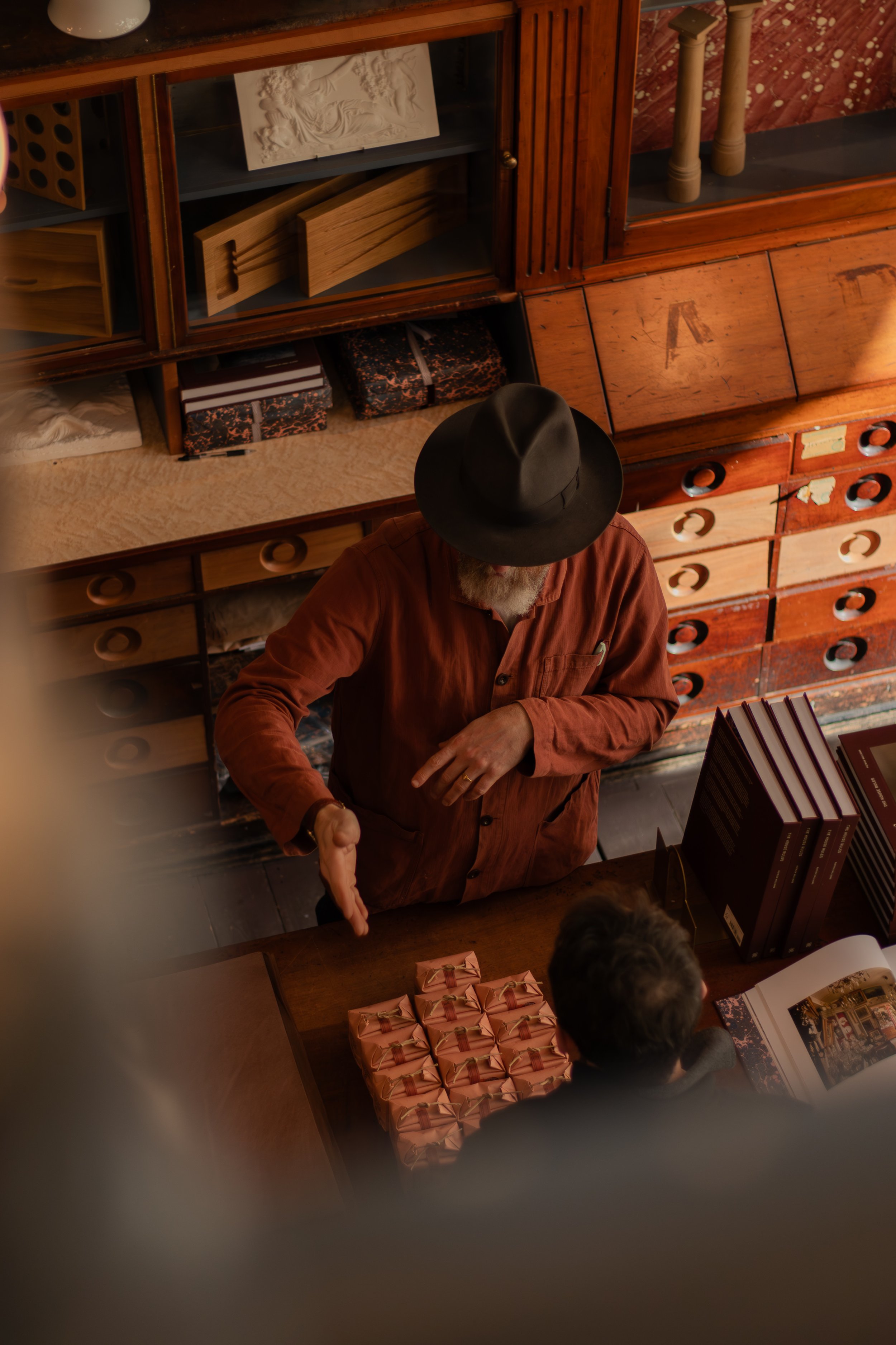 A man wearing a black hat and a brown shirt is extending his hand for a handshake across a table. The table has pink gift boxes tied with ribbons, and there is a person with short dark hair and glasses sitting across from him. The background includes