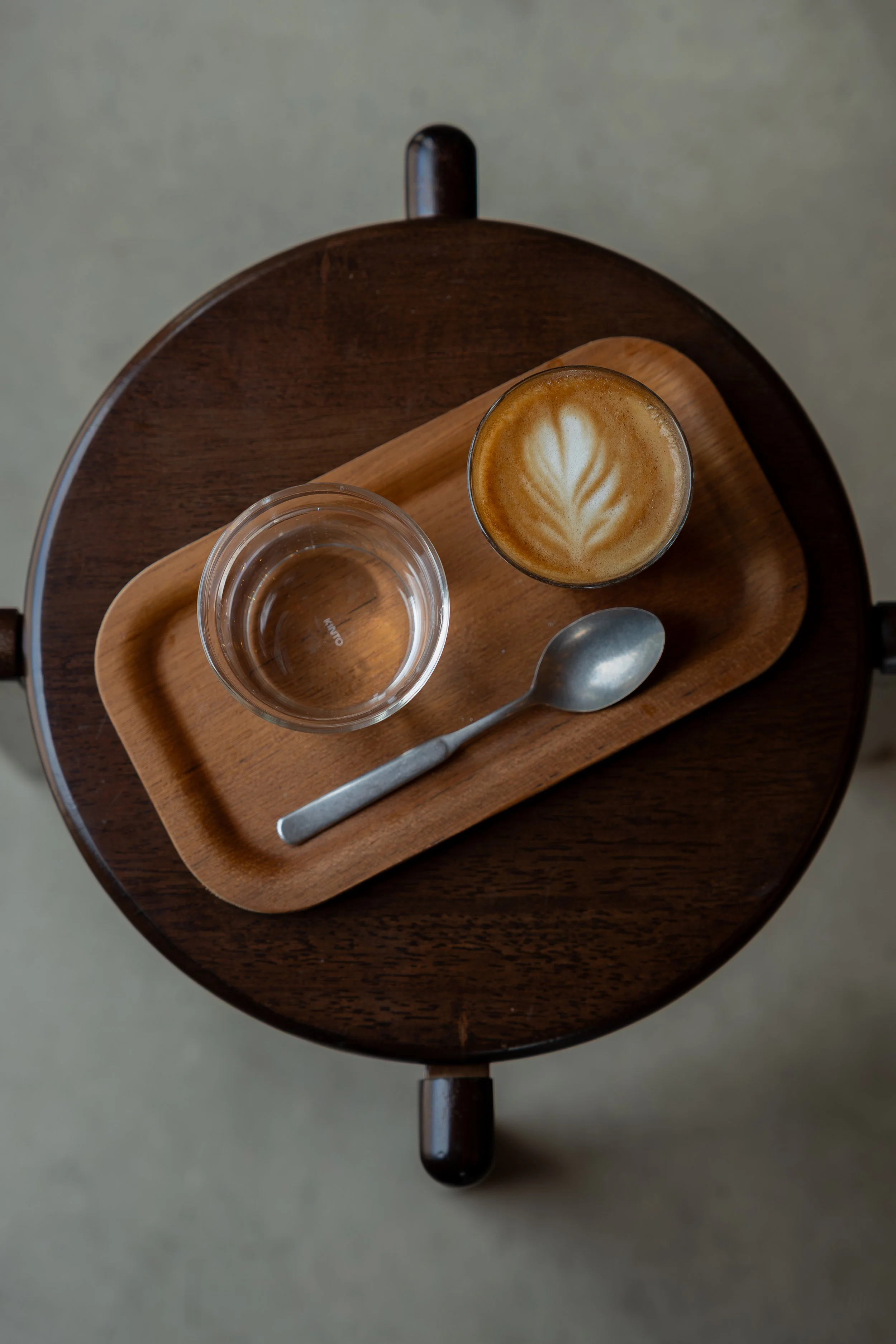 Top-down view of a wooden tray with a glass of water and a latte coffee with latte art on a round wooden table.