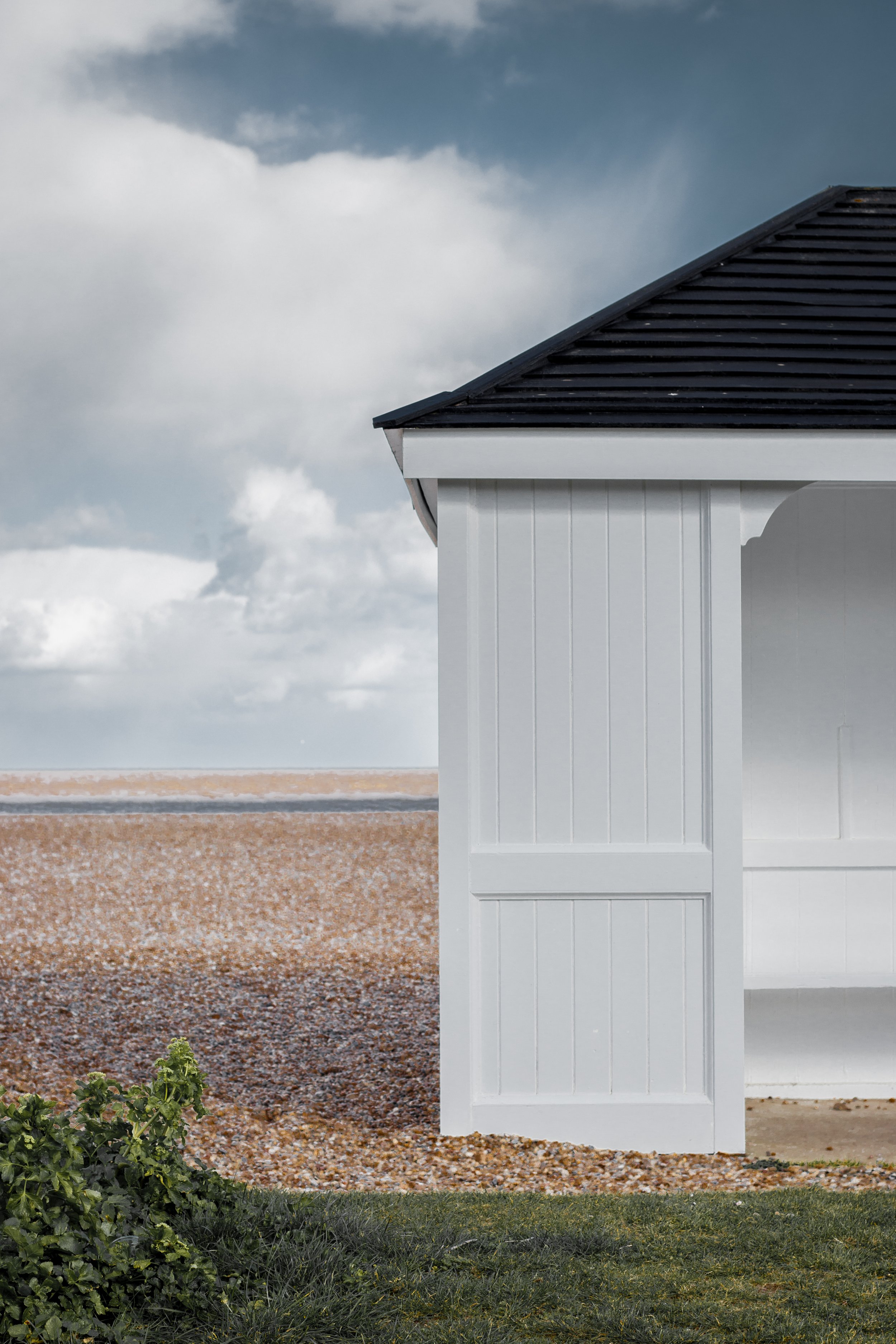 A white shed with vertically paneled walls and a black sloped roof, positioned near a grassy area and pebbled ground, with a background of cloudy sky in Aldeburgh, United Kingdom.
