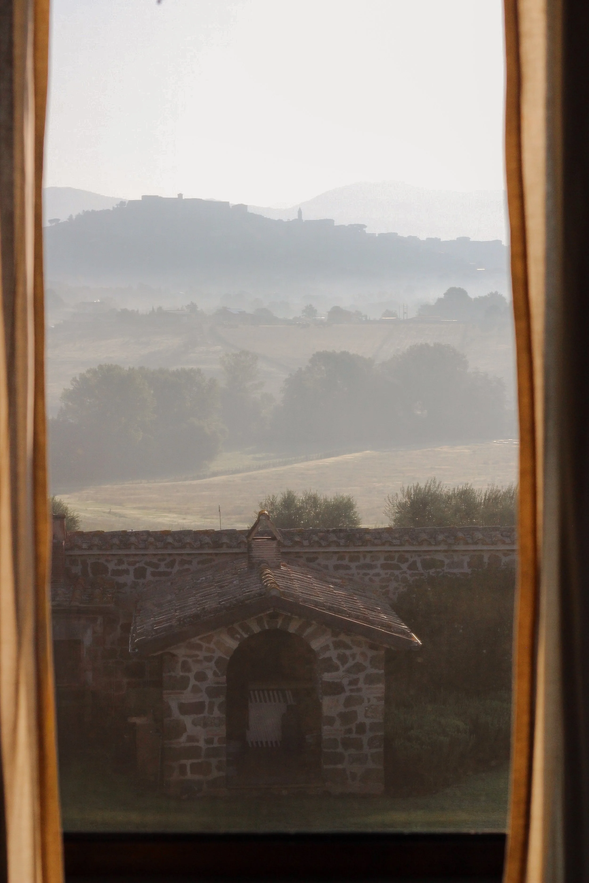 View of a hillside with buildings and trees in Italy, seen through a window with wooden curtains, a stone building with a tiled roof in the foreground.