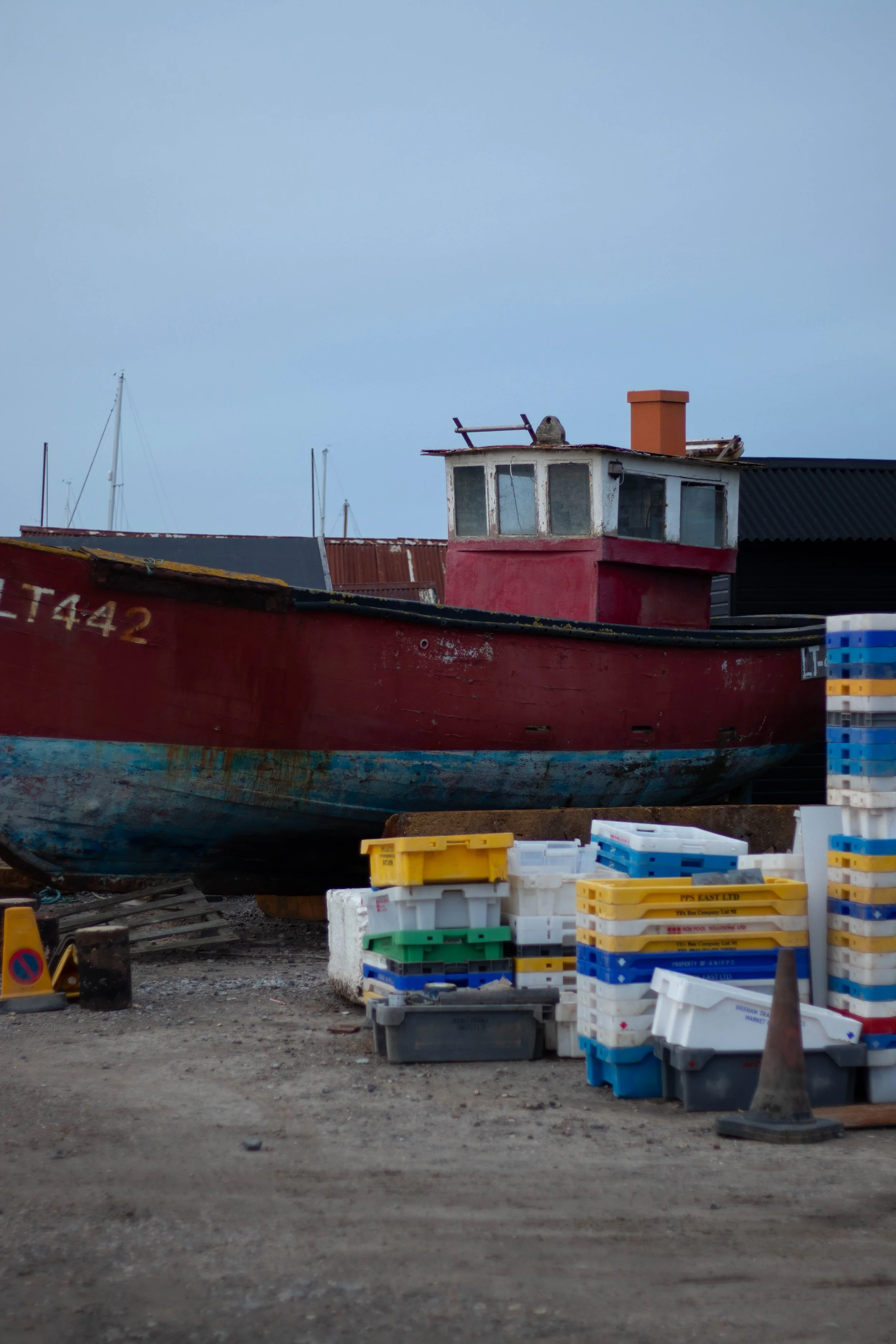 An old, red fishing boat with a worn hull parked on land near fishing crates and cones, with sailboats visible in the background in Southwold, United Kingdom.