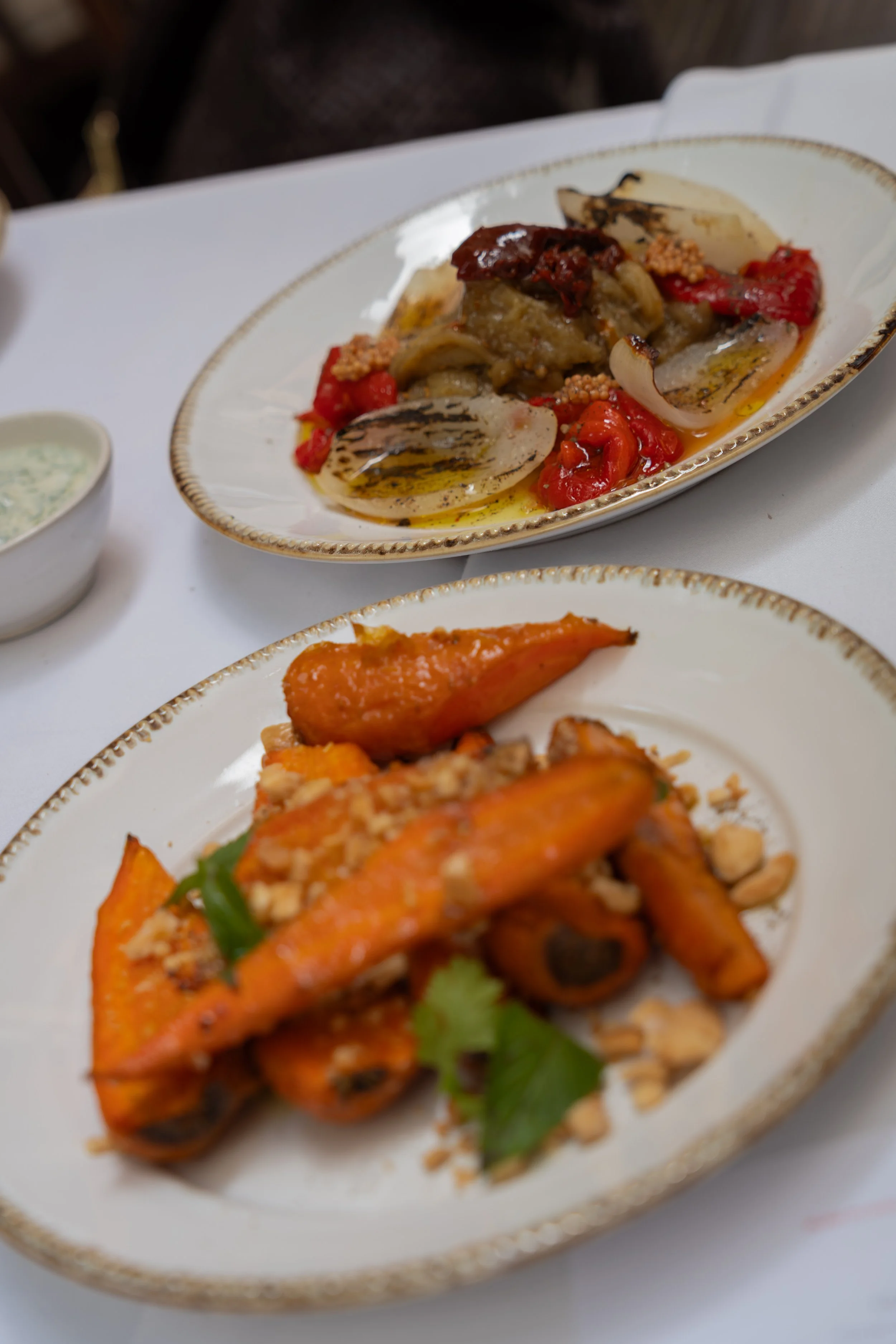 Plate of grilled vegetables, including eggplant, red bell peppers, and garlic, with a sauce, on a white tablecloth, with some other dishes partially visible at Coloma restaurant in Palma, Majorca.