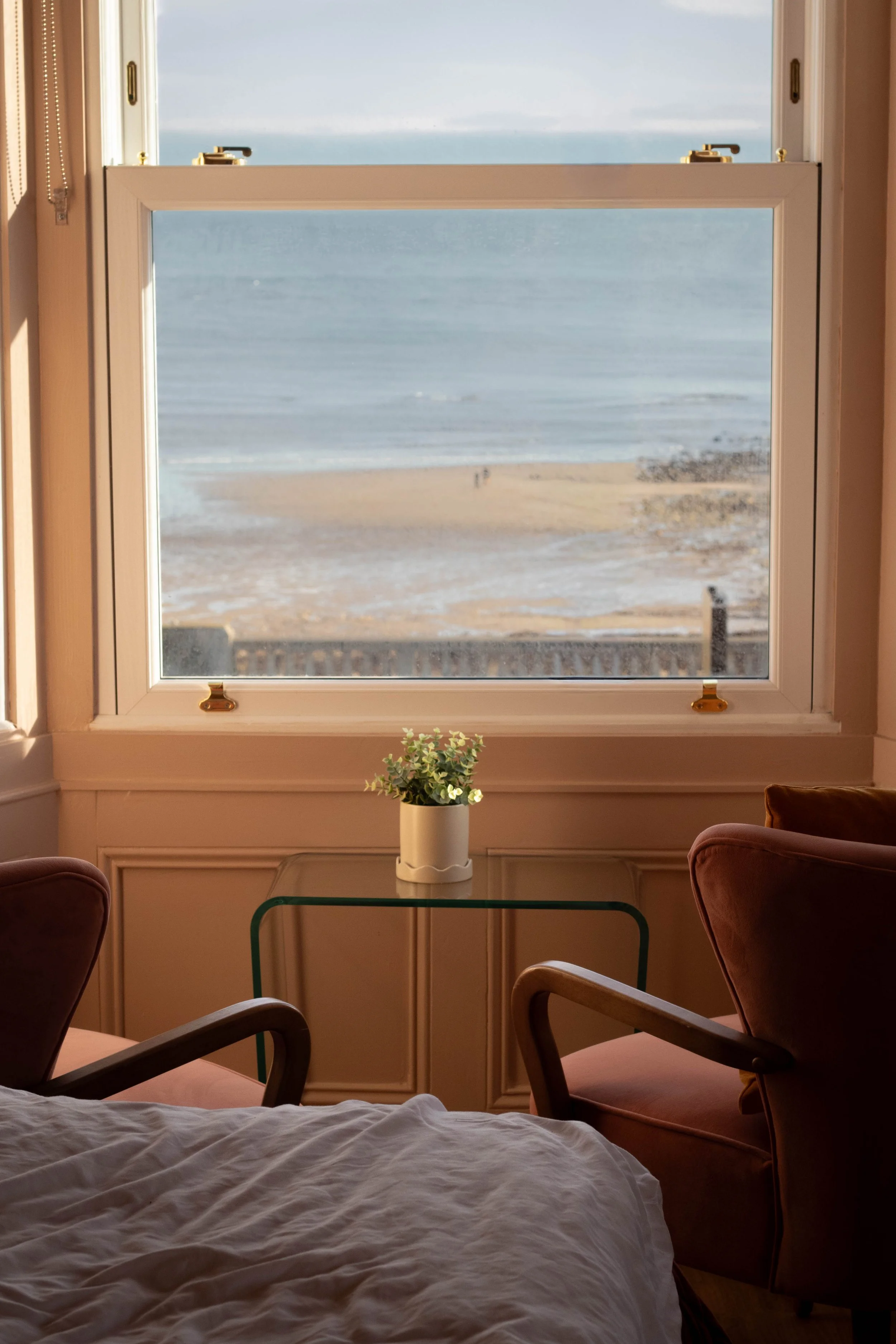 Room with a large window overlooking a beach and ocean, with two chairs and a small glass table holding a potted plant in Roker, United Kingdom.