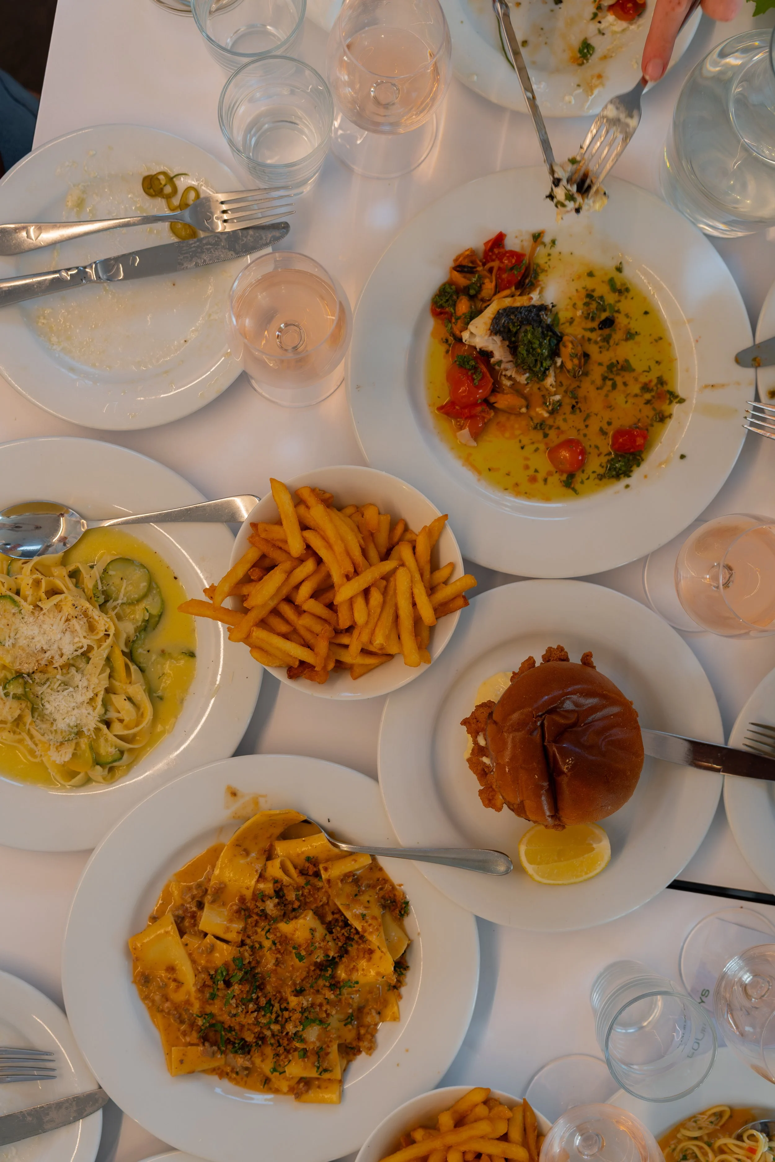 Top-down table view of empty plates with remnants of pasta, vegetables, and sauce, along with glasses of water, wine, and a burger with fries, on a white table at a meal gathering at Four Boys restaurant in Rock, Cornwall.
