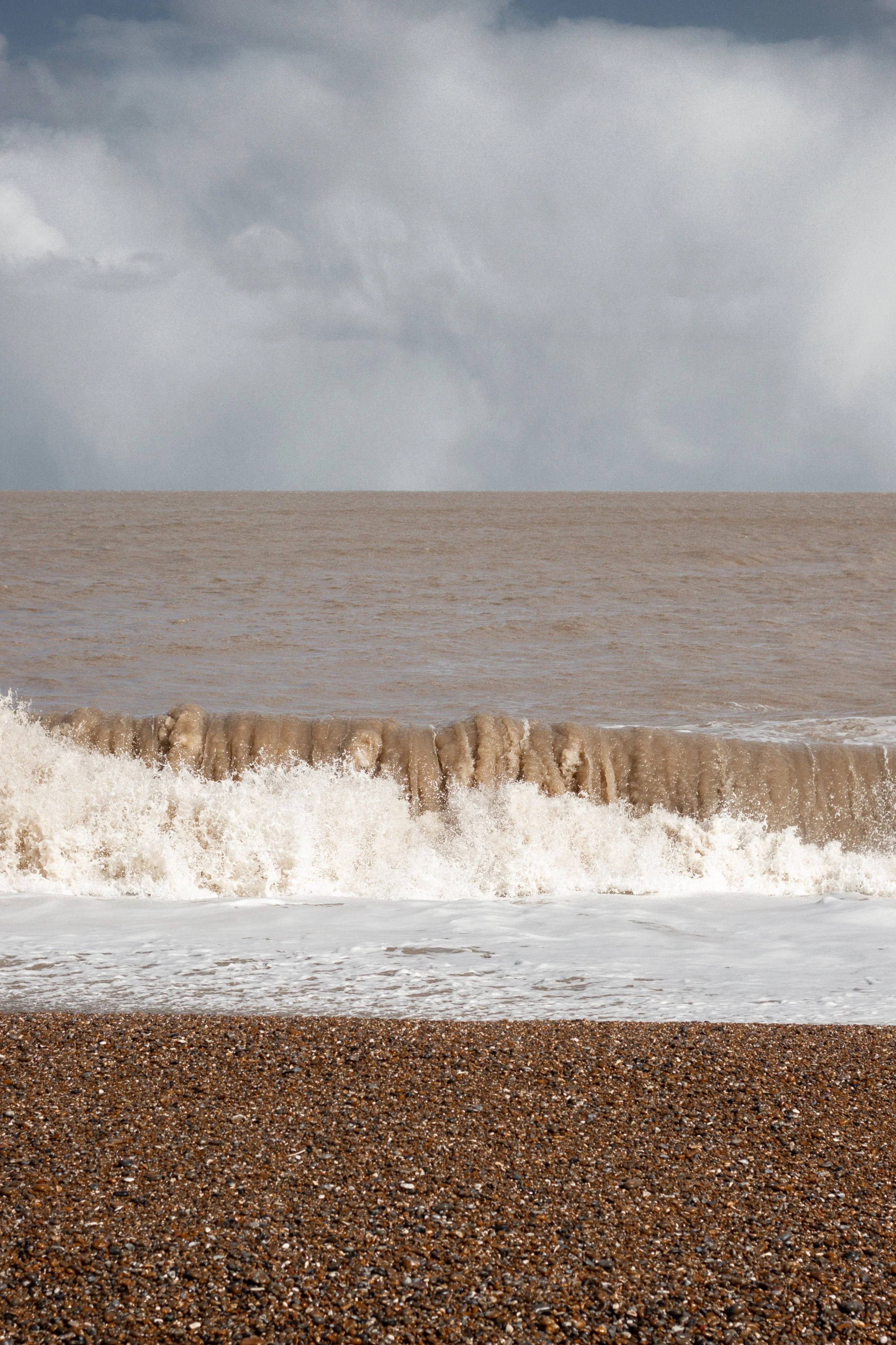Cloudy sky over a brownish ocean with waves crashing onto a pebbled beach in Aldeburgh, United Kingdom.