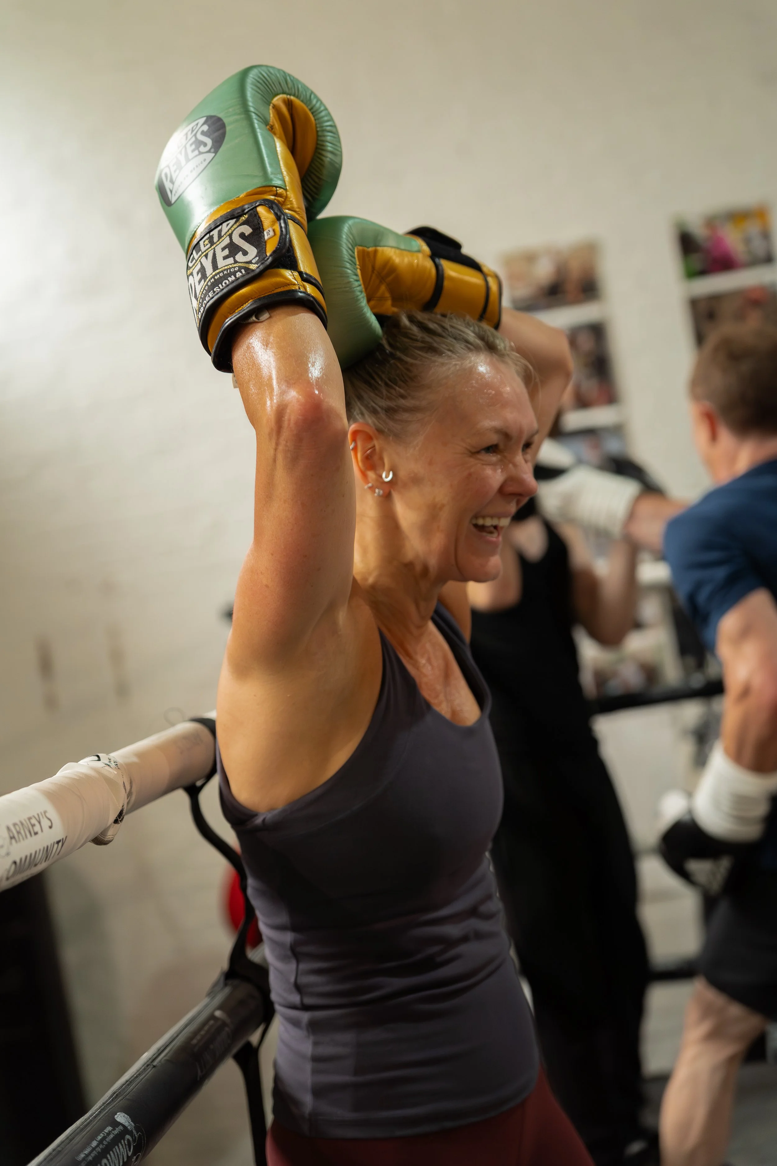 A woman with a broad smile, wearing a black athletic top and maroon shorts, standing in a boxing gym, holding green boxing gloves over her head.