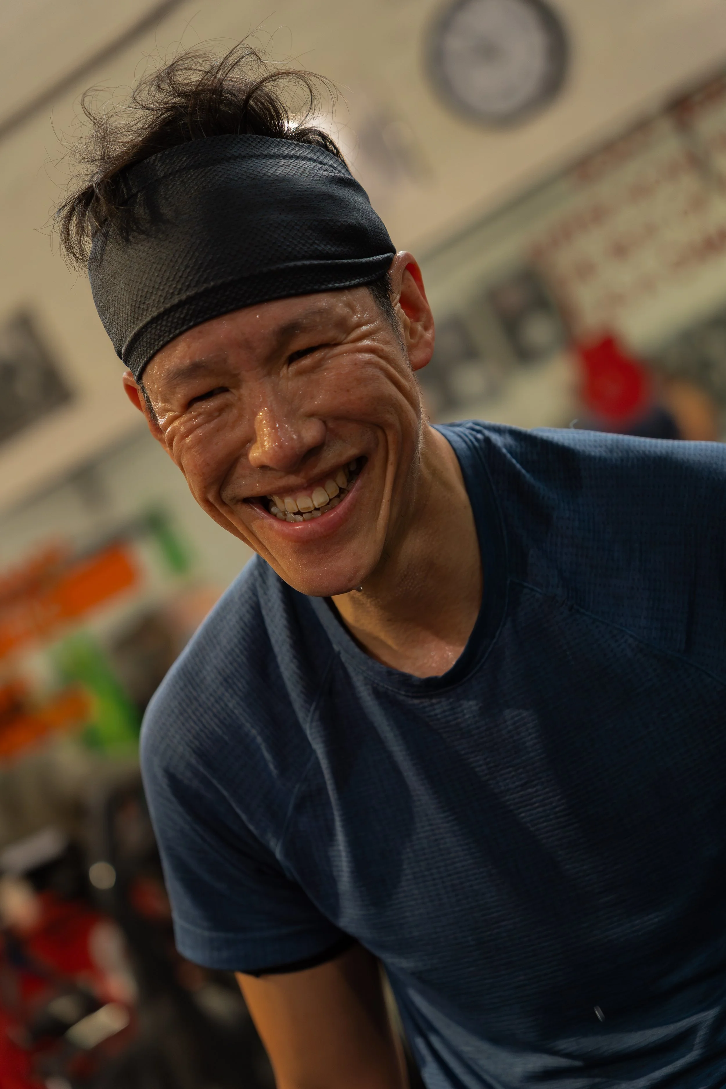 A man with short dark hair and a black headband smiling and looking down, wearing a dark blue shirt, in a gym with blurred equipment in the background.