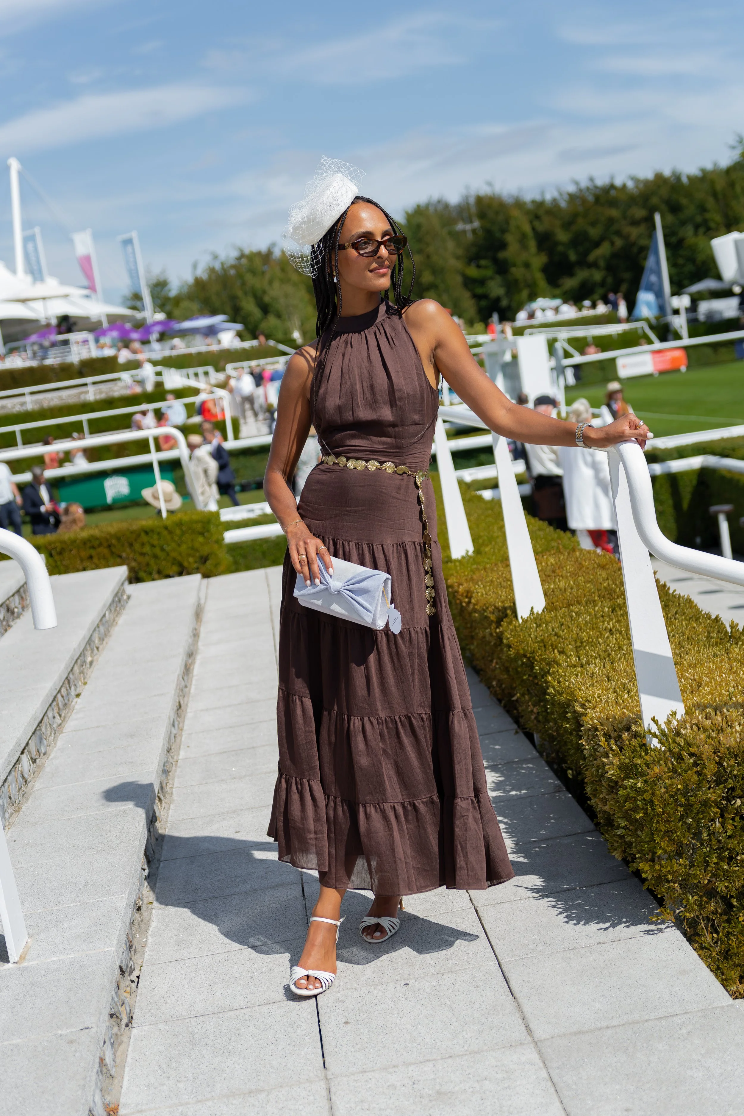 A woman in a long brown Phase Eight dress with a wide-brimmed hat and sunglasses walking outdoors near a railing, with a dressage arena and spectators in the background on a sunny day at Goodwood Racecourse.