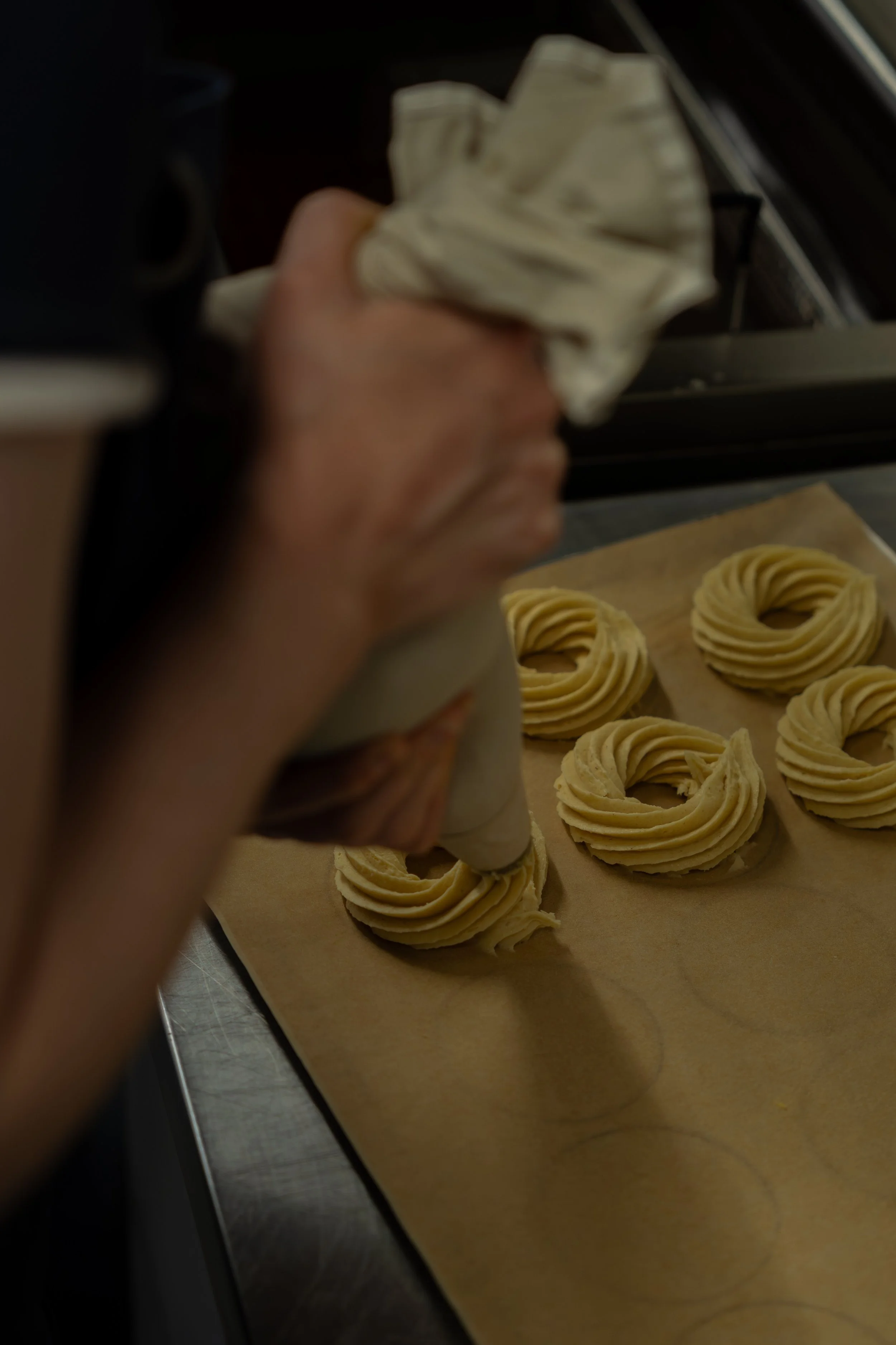 Close-up of a person squeezing pastry cream into rosette piping on parchment paper with un-fried cruller dough rounds