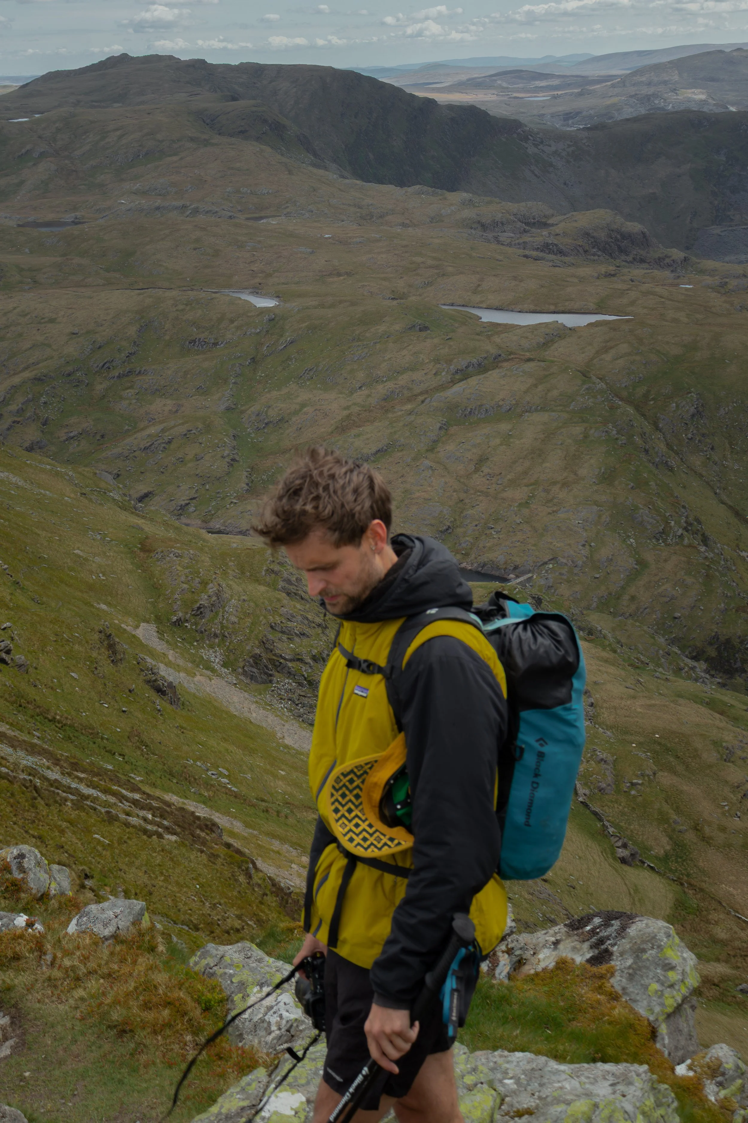 A young man wearing a yellow and black jacket and carrying a blue backpack hiking through a mountainous landscape with green grass, rocks, and small lakes, overcast sky.