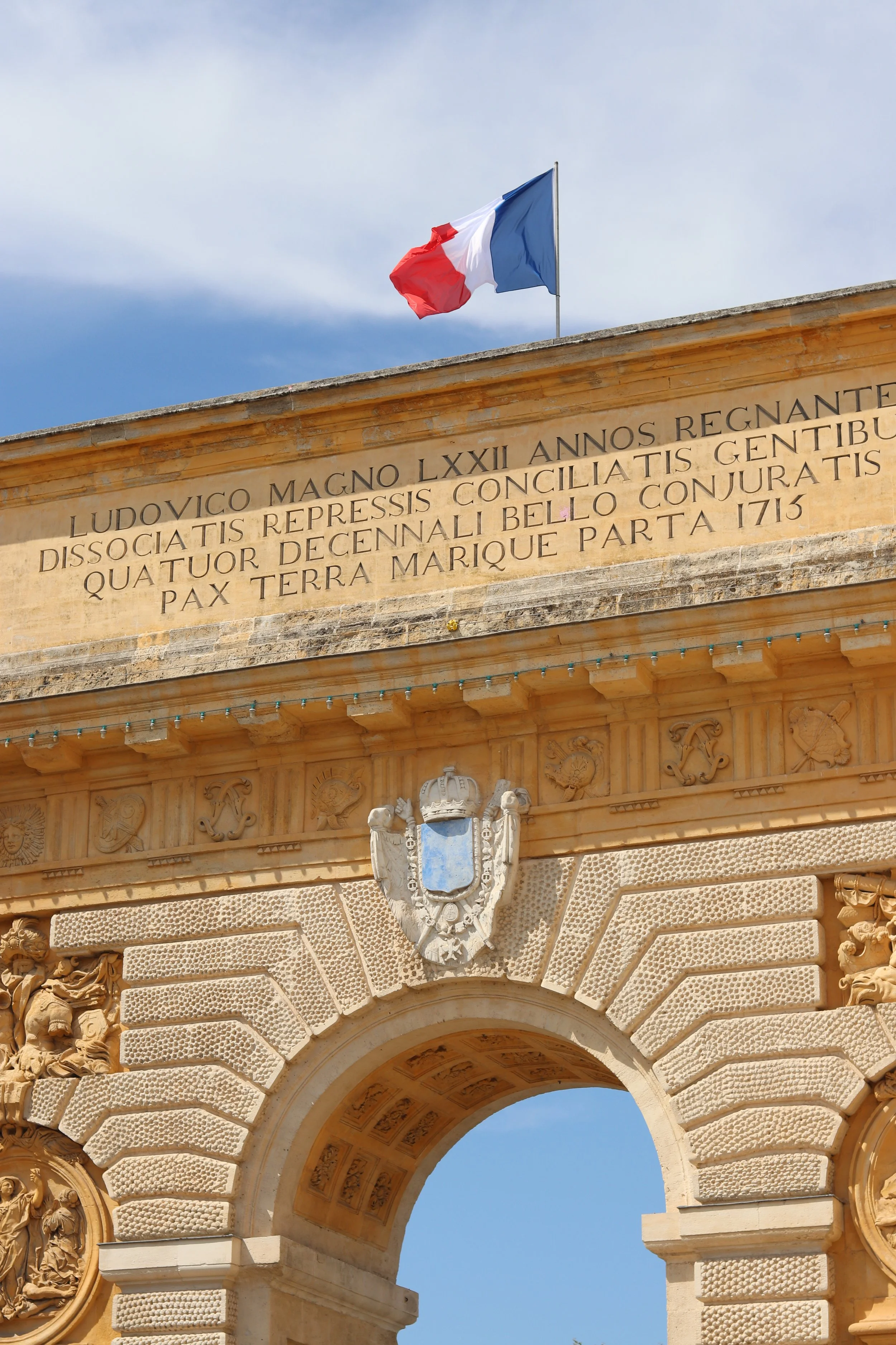 A historic arch with an inscription in Latin, the French flag on top, and a blue sky in the background.