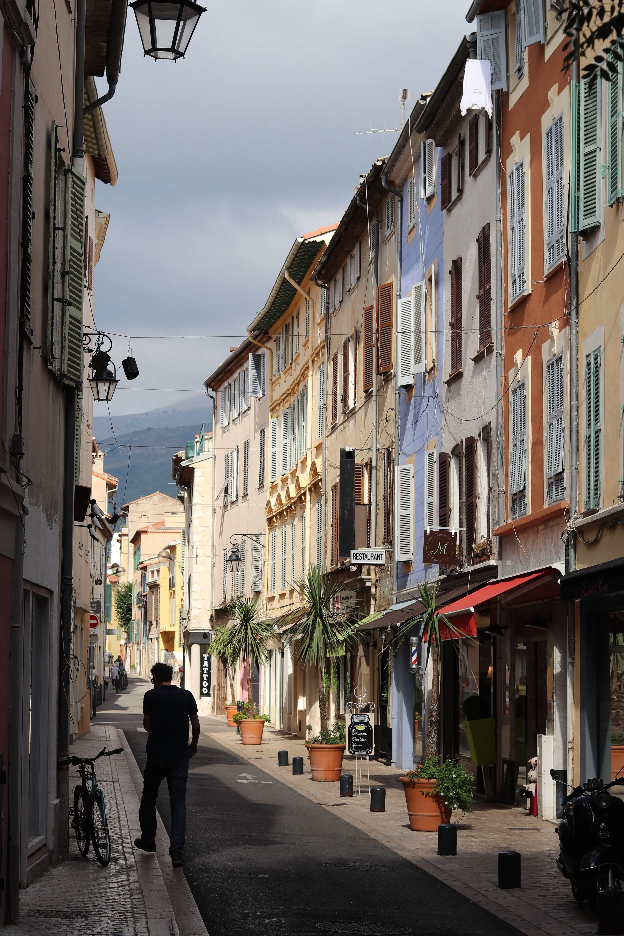 A narrow street in a French town with colourful buildings and shuttered windows. Potted palm trees and storefronts line the sidewalk. A person walks away from the camera, and bicycles and a scooter are parked along the street. The sky is overcast.