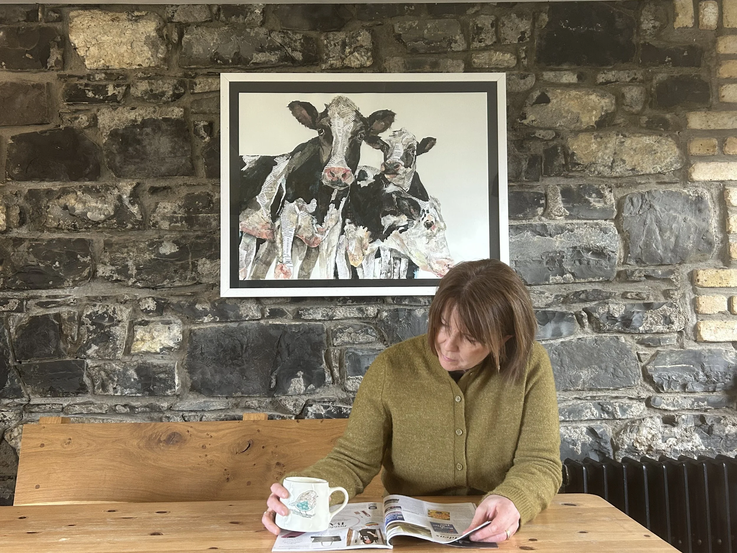 A woman with brown hair wearing a green cardigan is sitting at a wooden table, reading a magazine and holding a cup with a landscape design. Behind her, there is a stone wall with a framed painting of two cows, one black and white and the other mostly white with black patches.