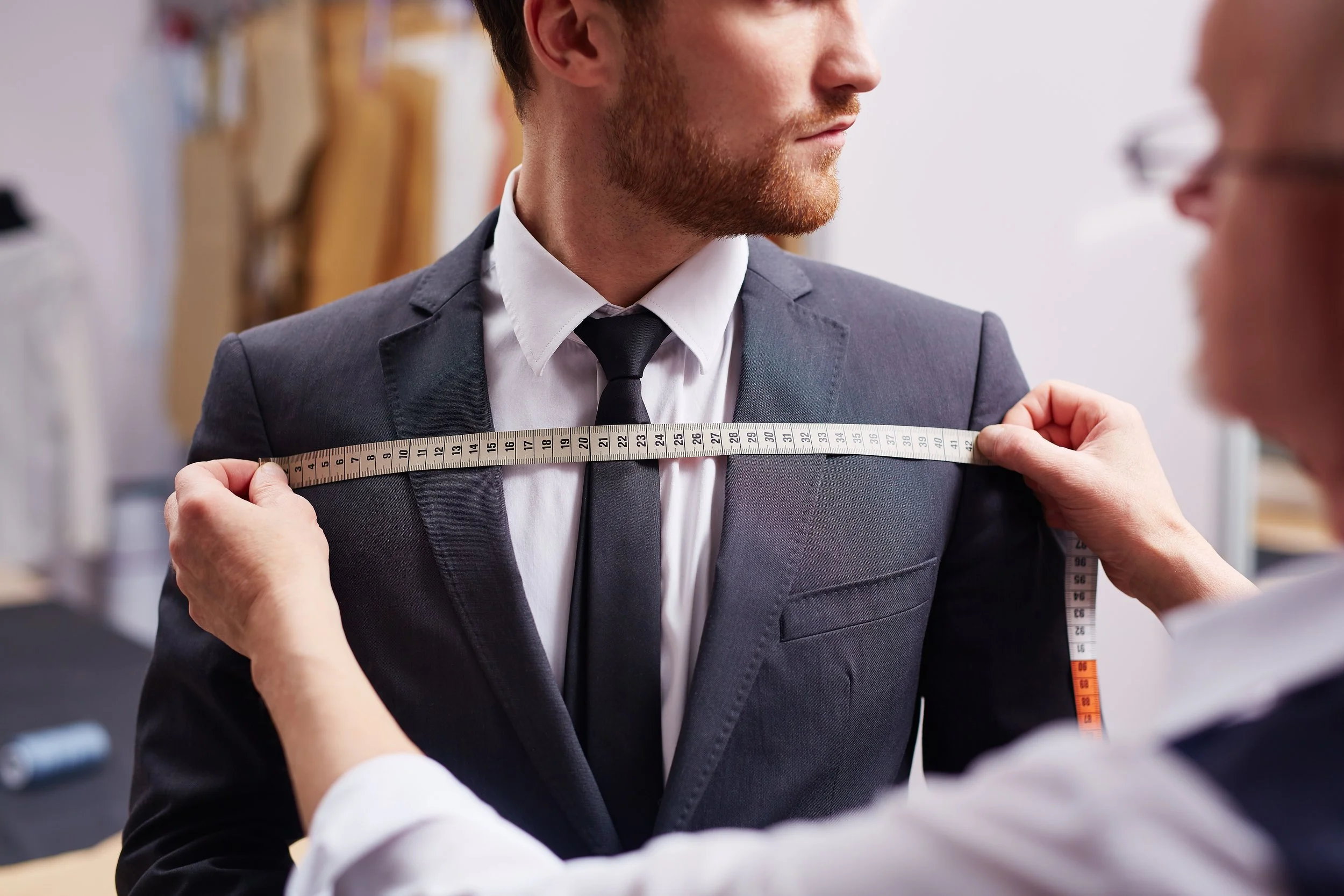 A man in a suit is being measured for his chest with a measuring tape in a clothing fitting room.