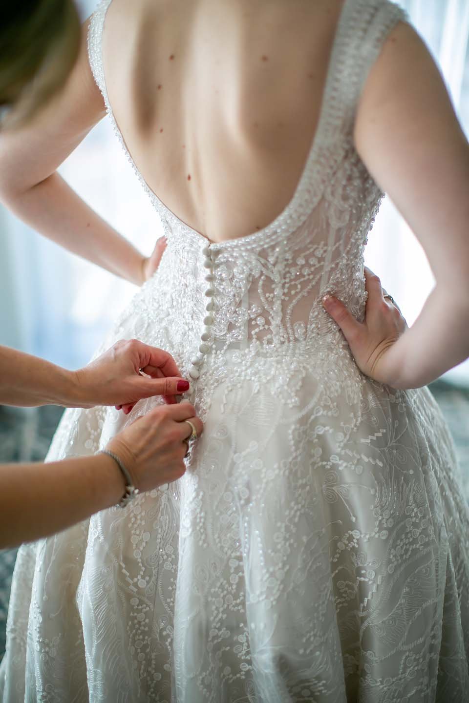 A woman is being helped into a wedding dress by another person, with the dress featuring intricate lace and beaded details, and buttons being fastened down the back.