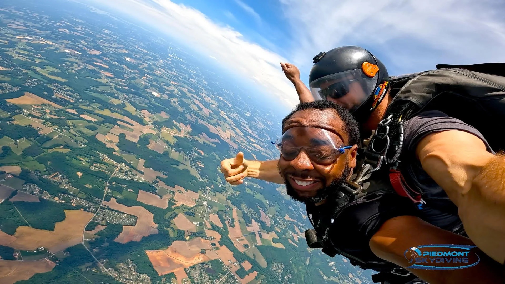 Two skydivers smiling and taking a selfie during free fall over a patchwork of farmland and countryside, with a partly cloudy sky above.