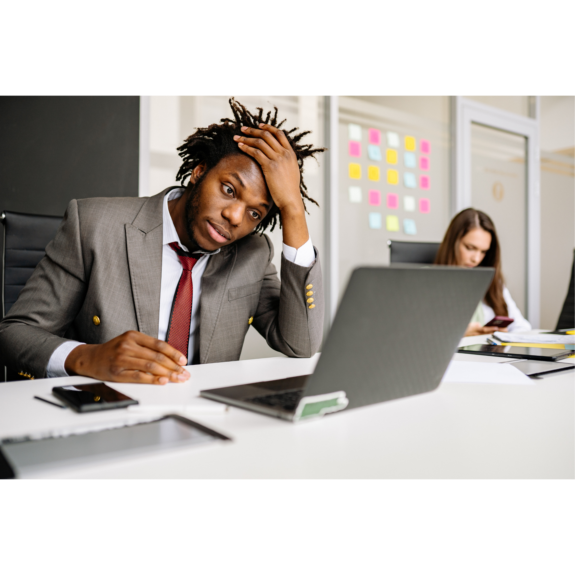 Man in a suit looking stressed at his desk in an office, with a laptop, smartphones, and papers, with a woman in the background using a smartphone.