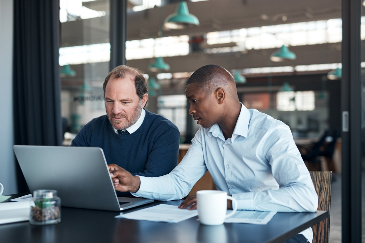 Two men sitting at a table, looking at a laptop together in a modern office or cafe setting.