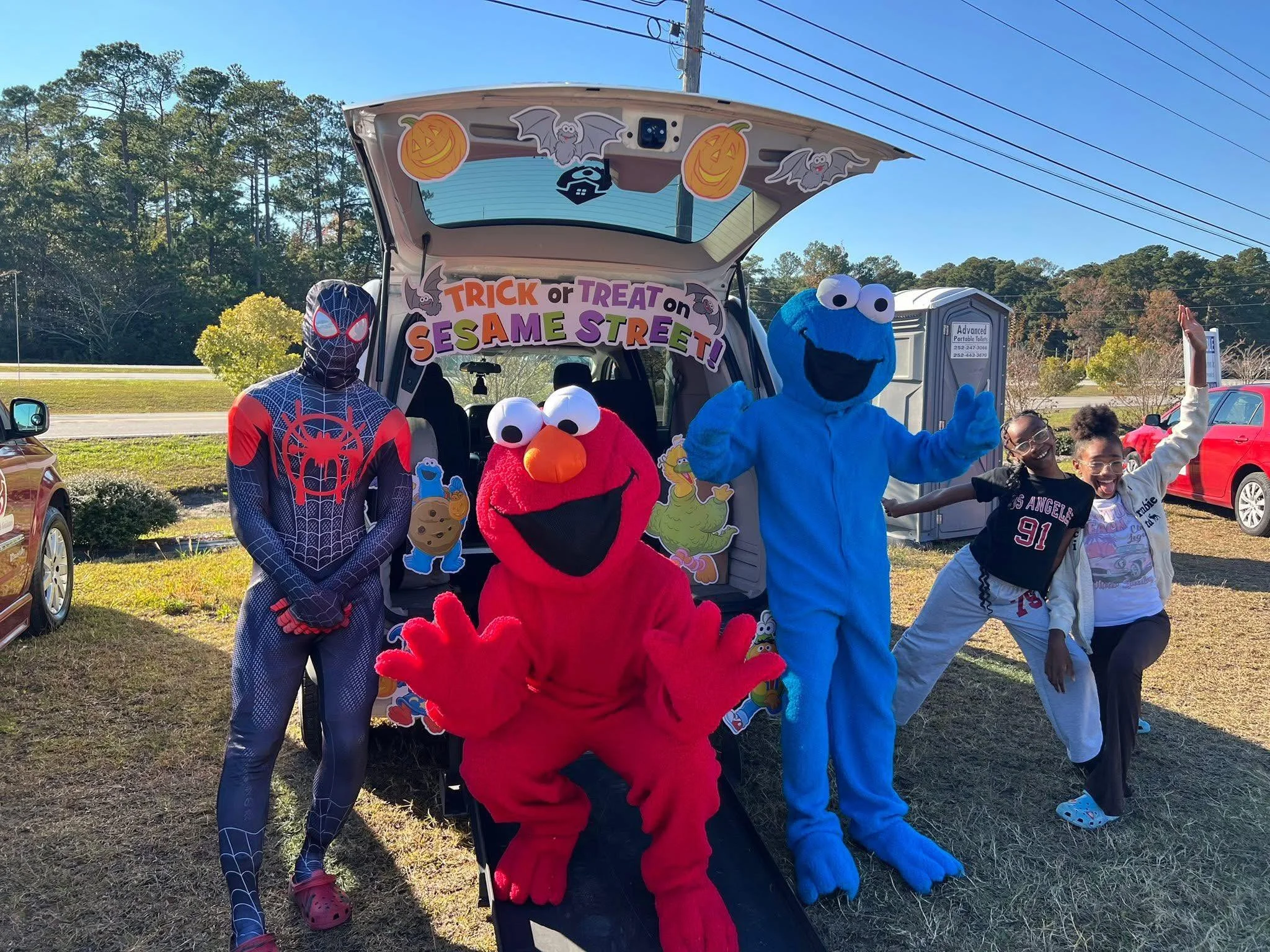 People dressed as Spider-Man, Elmo, Cookie Monster, and Sesame Street characters posing for a photo at a Halloween event with a truck decorated for Sesame Street and children smiling and making poses.